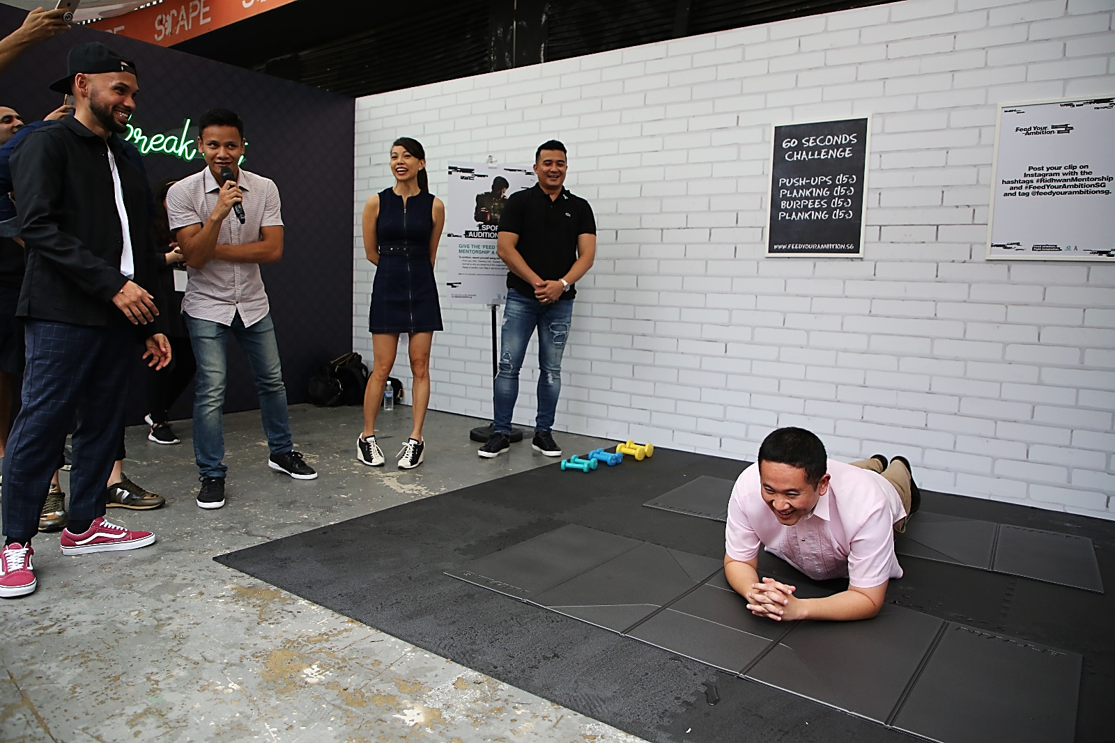 Several people are standing or watching a person planking on mats in front of a white brick wall with challenge instructions.