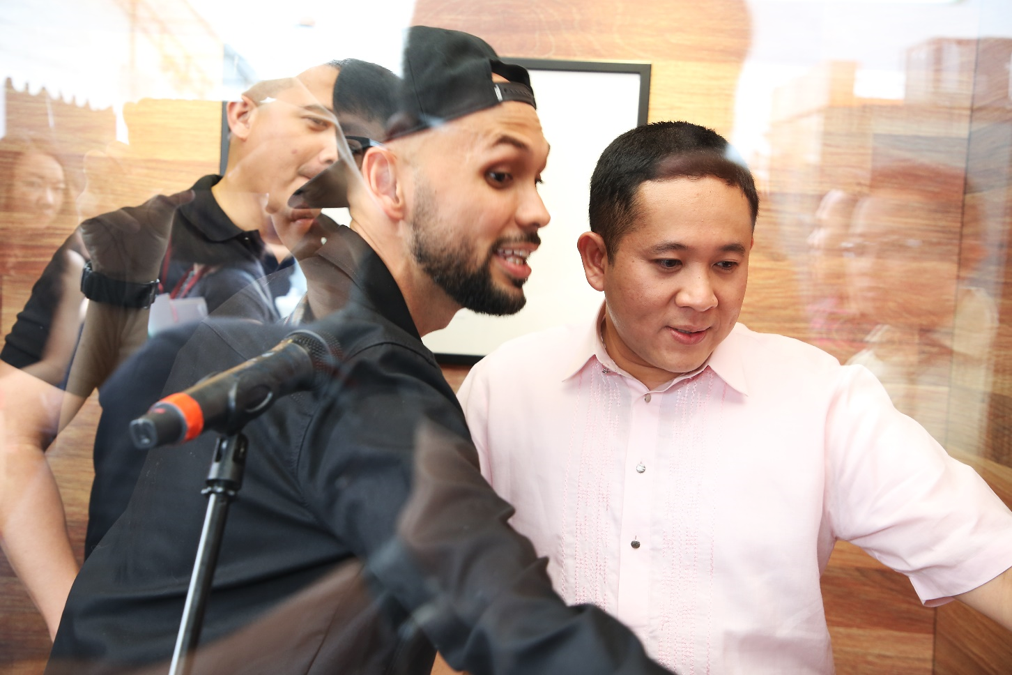 Two people are standing close to a microphone in a room with wooden walls and a framed picture.
