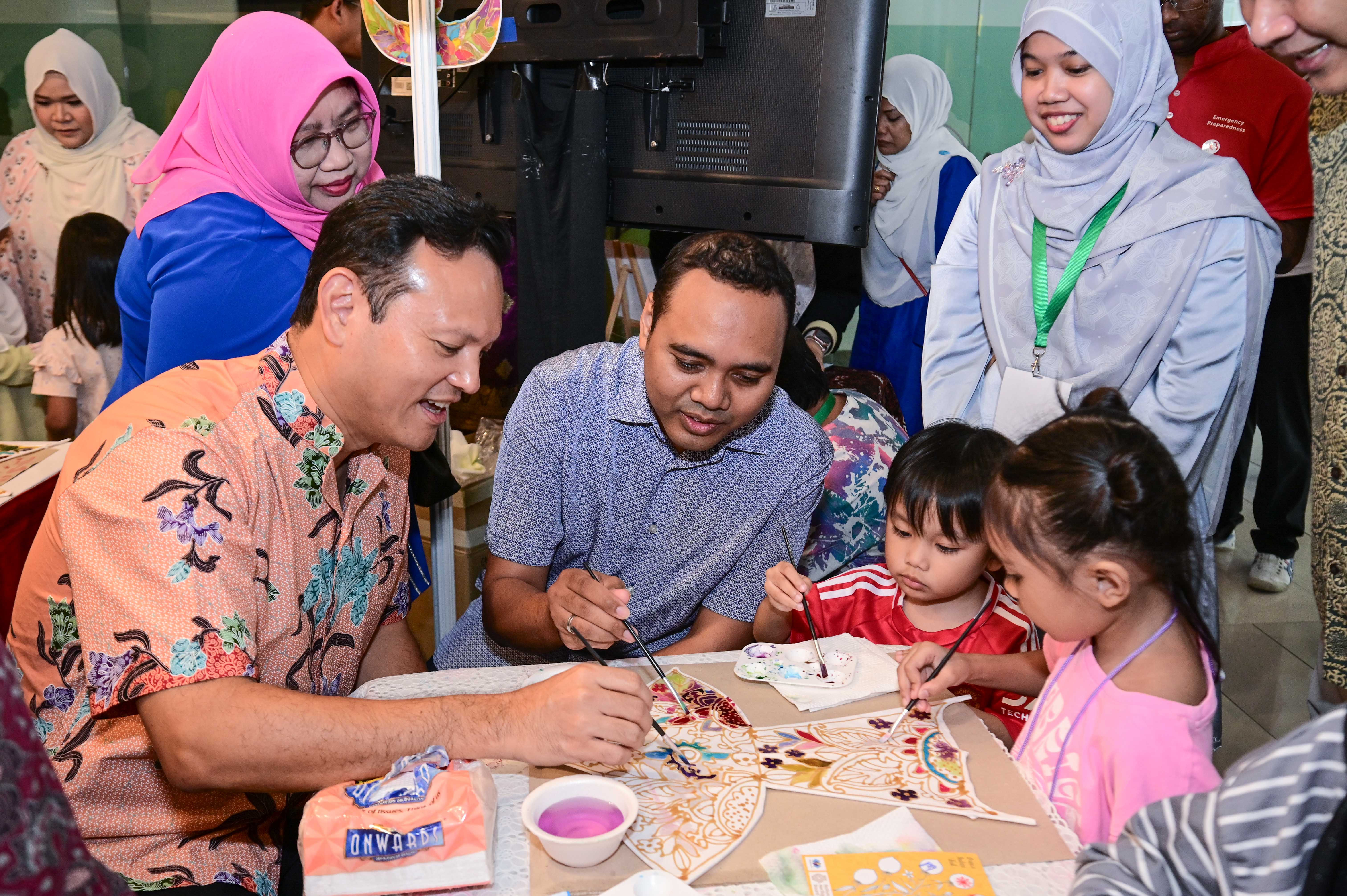 Adults and children are painting decorative fans at a crowded indoor event, with various people standing and watching nearby.