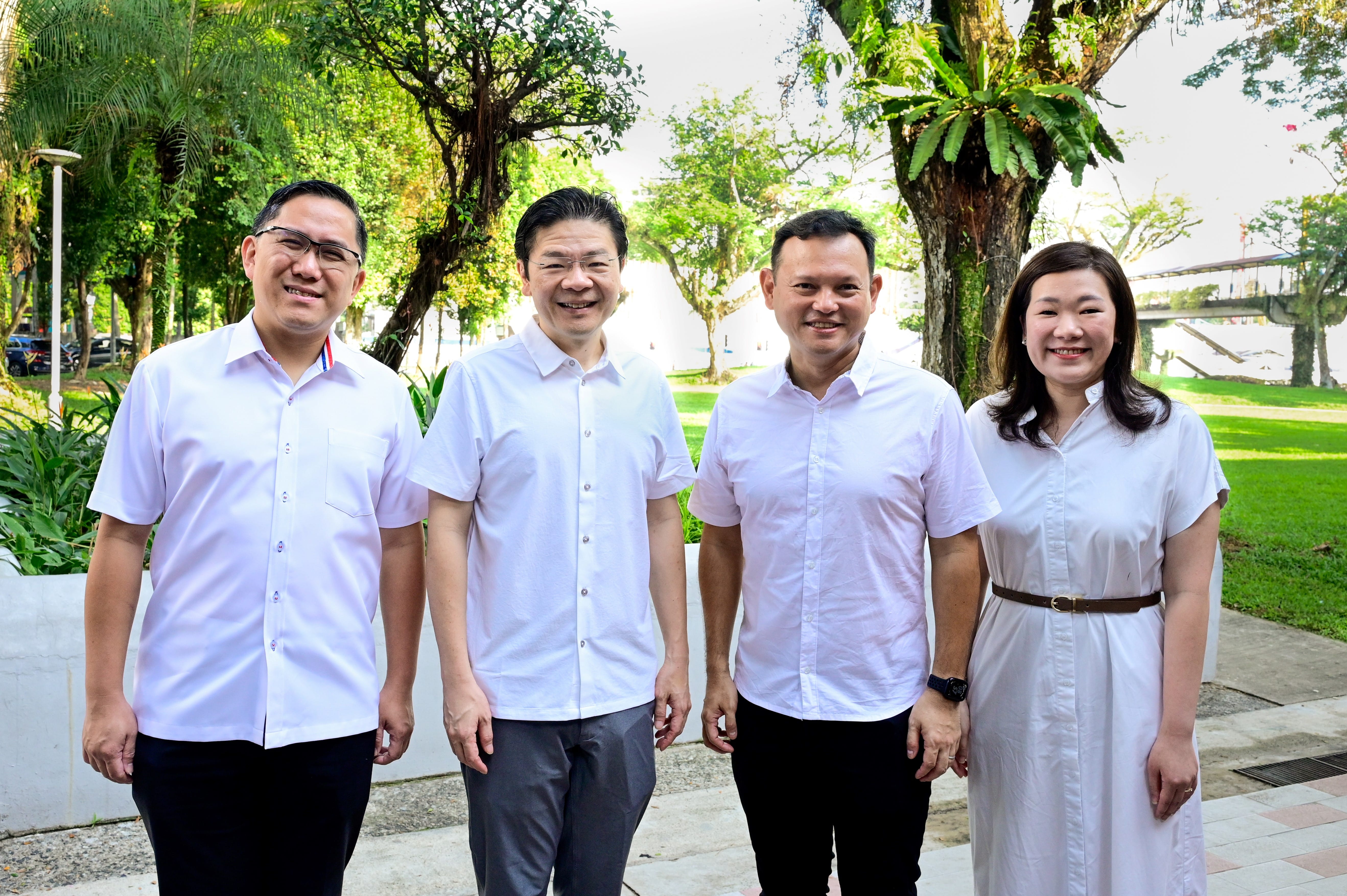 Four people stand outdoors wearing white shirts with greenery in the background.
