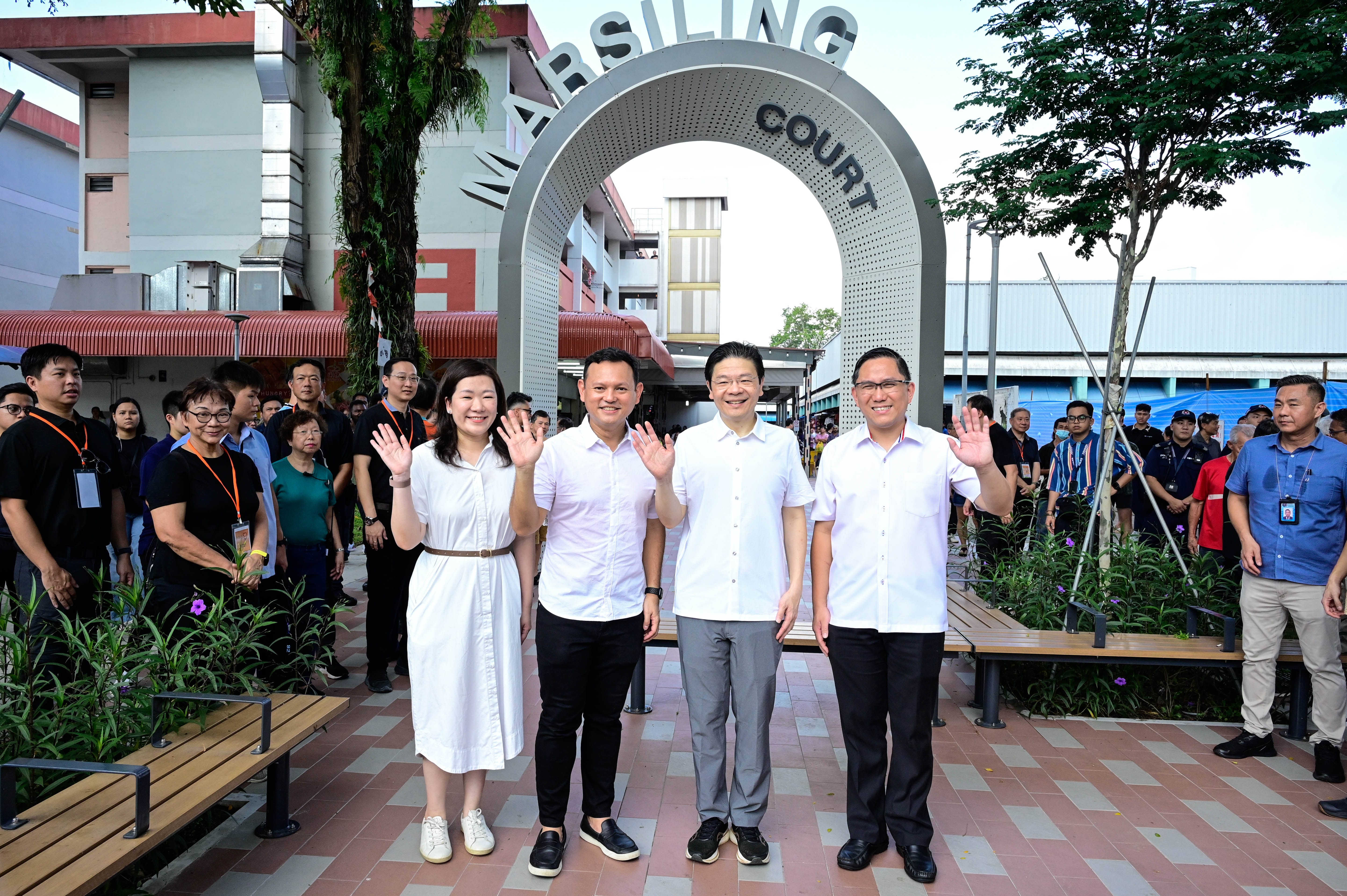 A group of people waving in front of an arch labeled "Marsiling Court" surrounded by buildings and greenery.