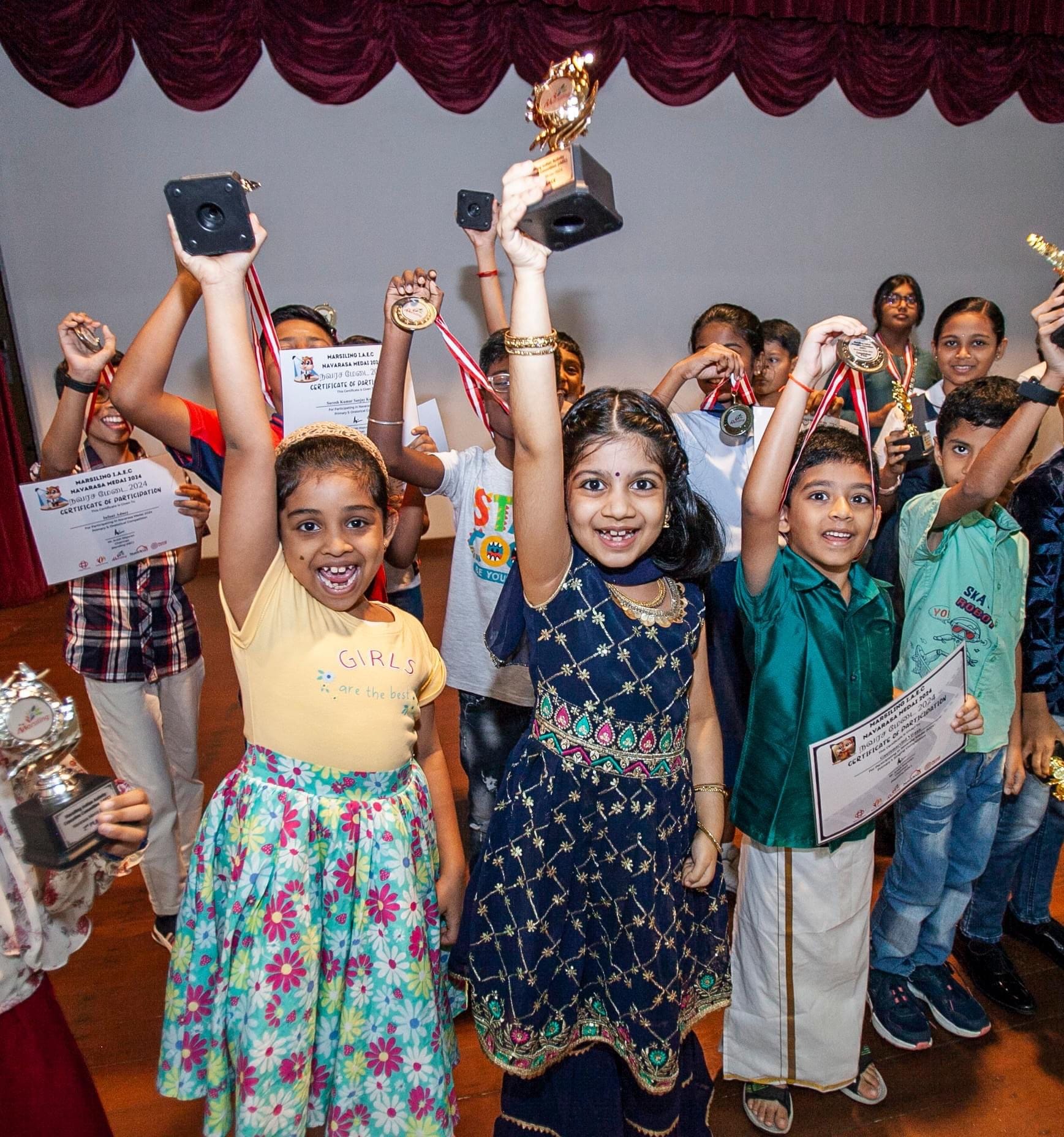 Children holding trophies, medals, and certificates while celebrating in a group on a stage.