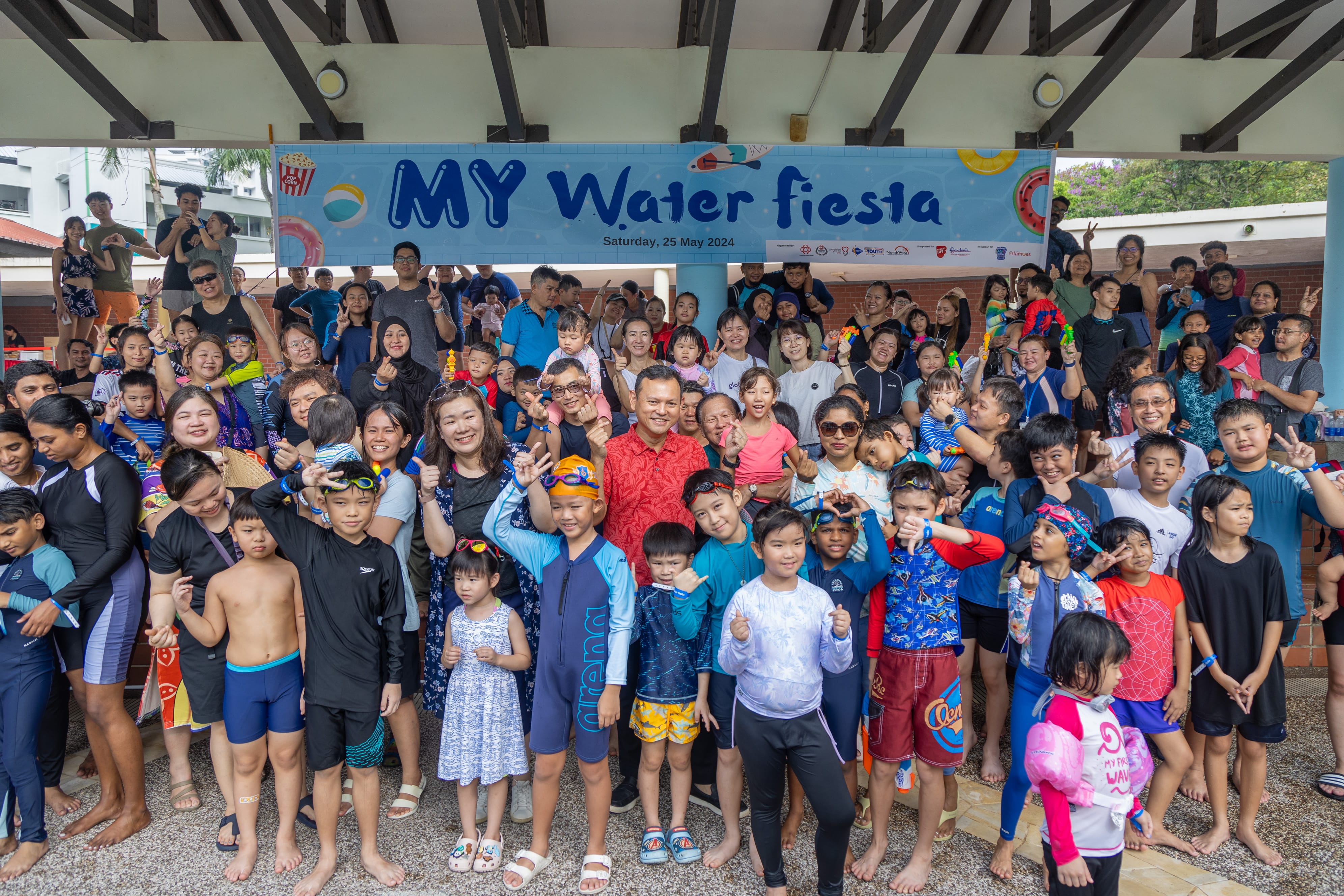 A large group of people, mostly children in swimsuits, are gathered under a banner that reads "MY Water Fiesta" dated 25 May 2024.