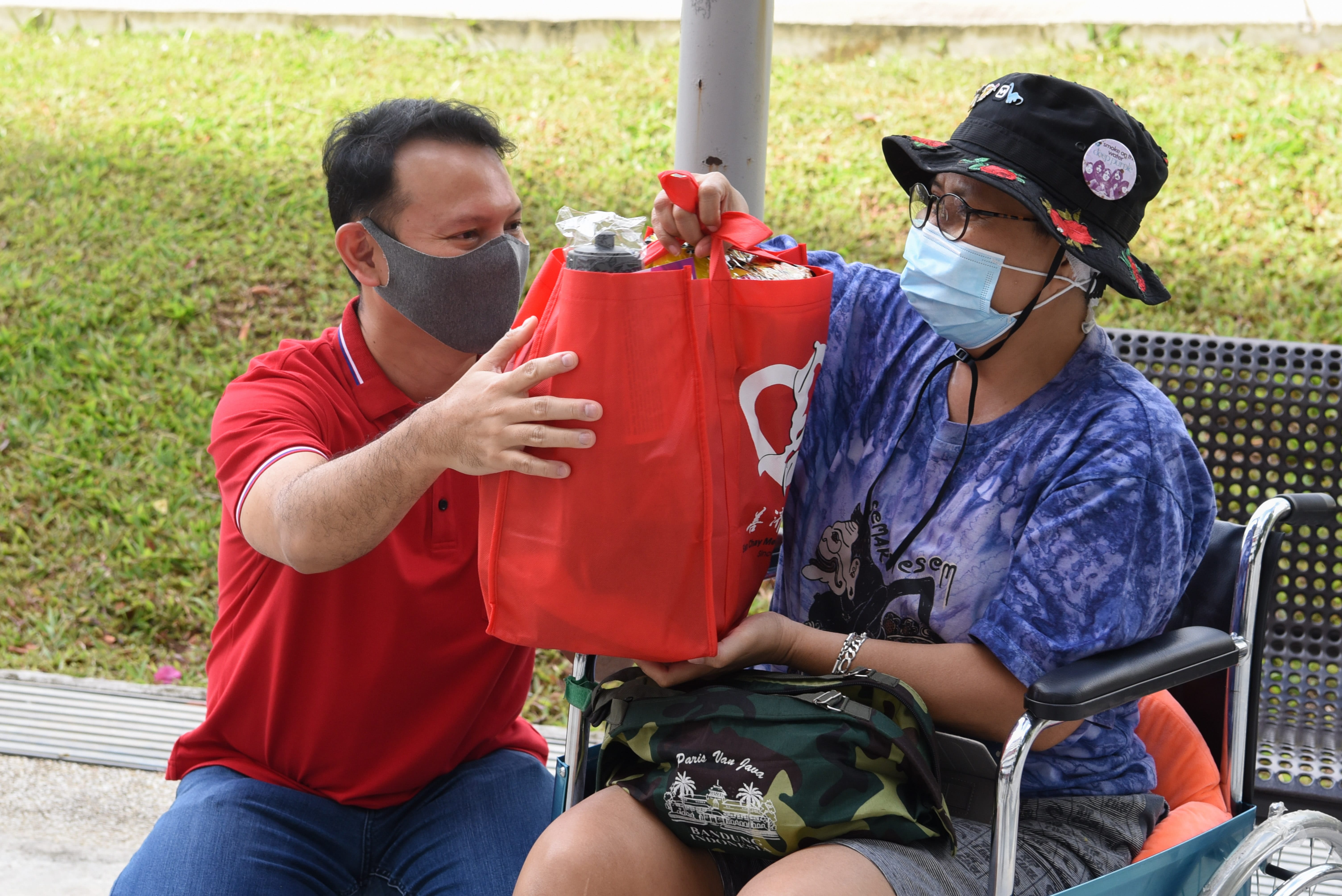 Masked man in red shirt hands a red tote bag of items to a masked woman in a wheelchair.
