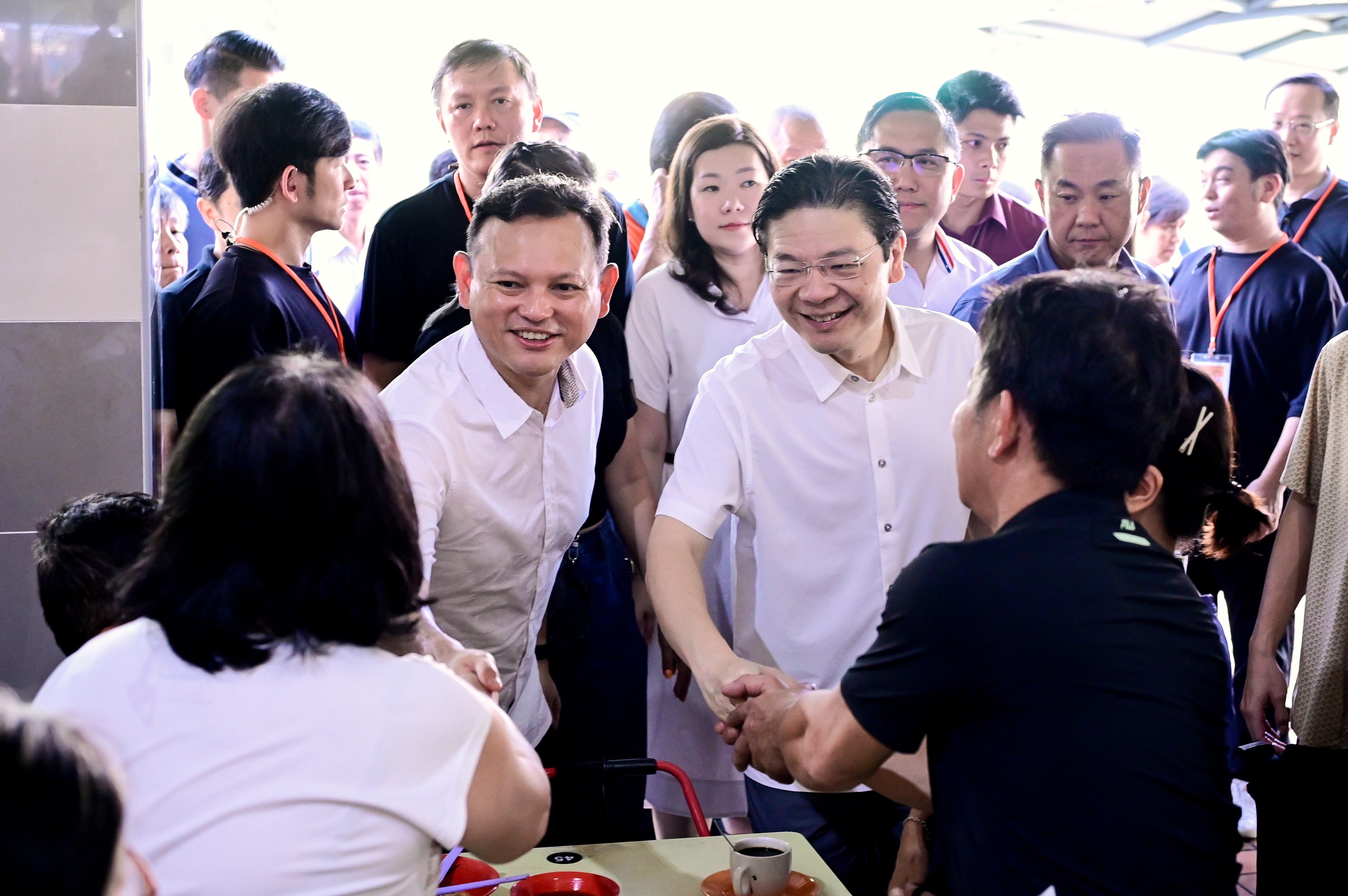 A group of people, one wearing a black shirt and another wearing a white shirt, engaging in a handshake in a crowded room.