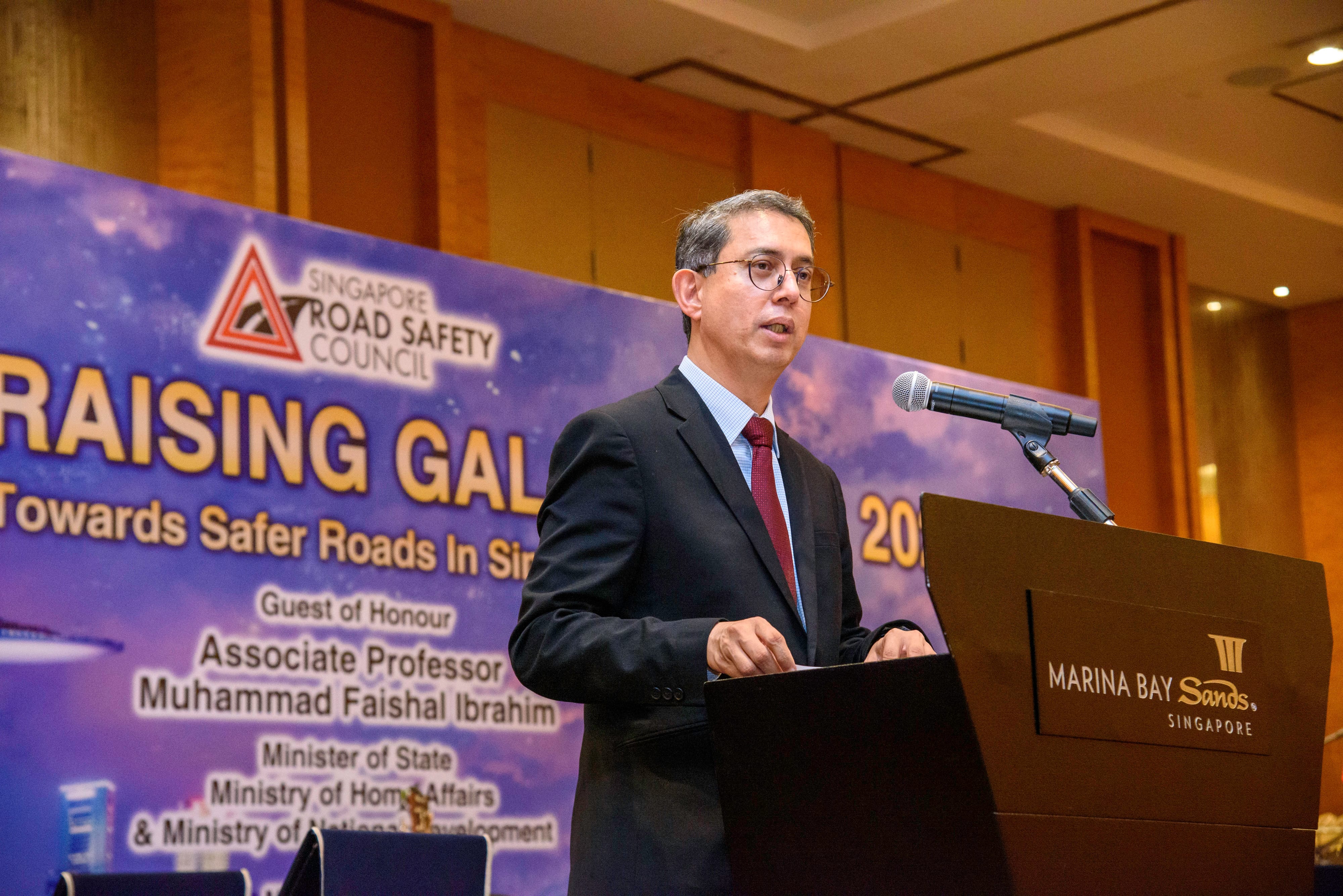 A man in a suit speaks at a podium with a microphone at the Singapore Road Safety Council event at Marina Bay Sands.
