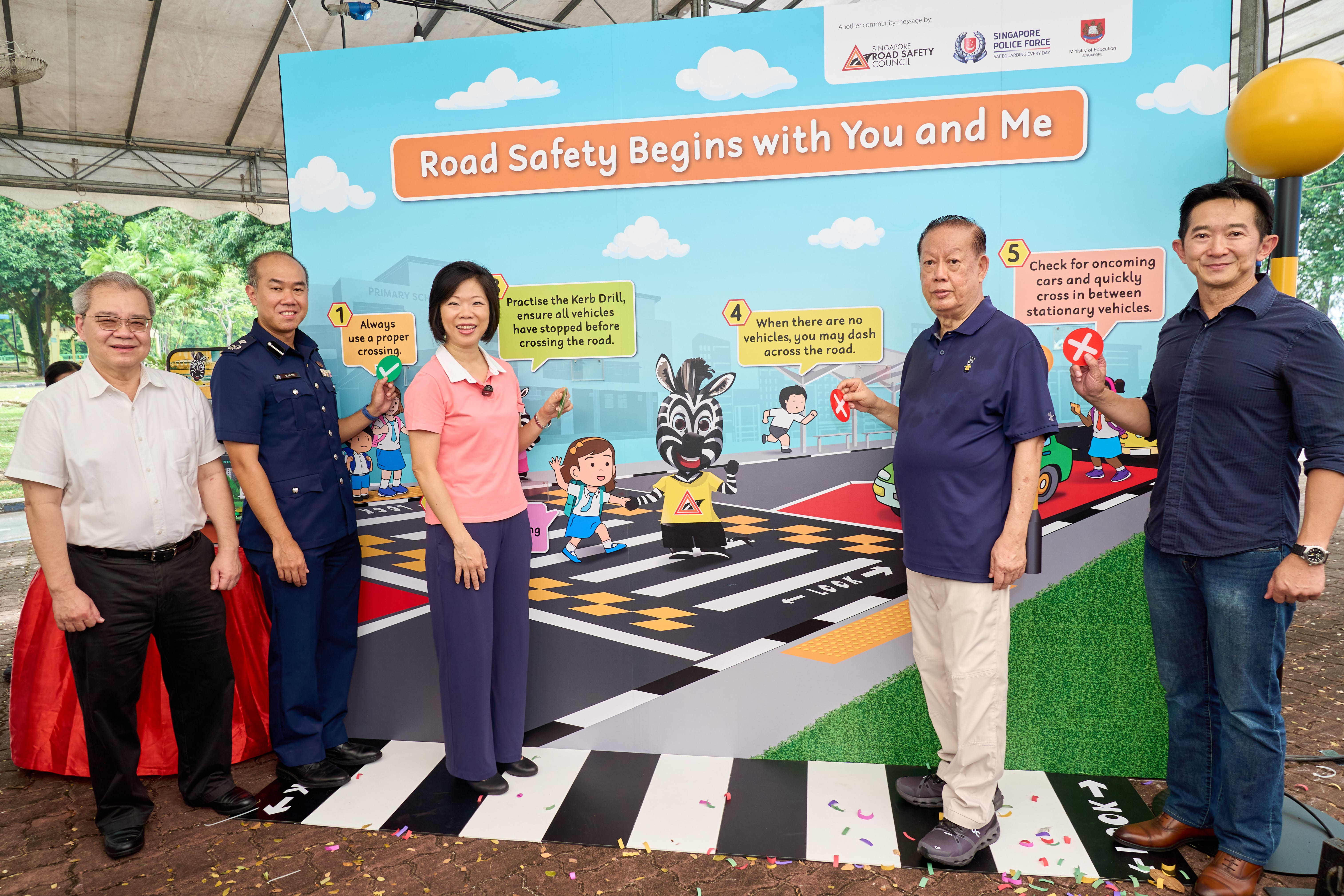 Five individuals standing in front of a large display about road safety measures with illustrated children and zebra mascot.