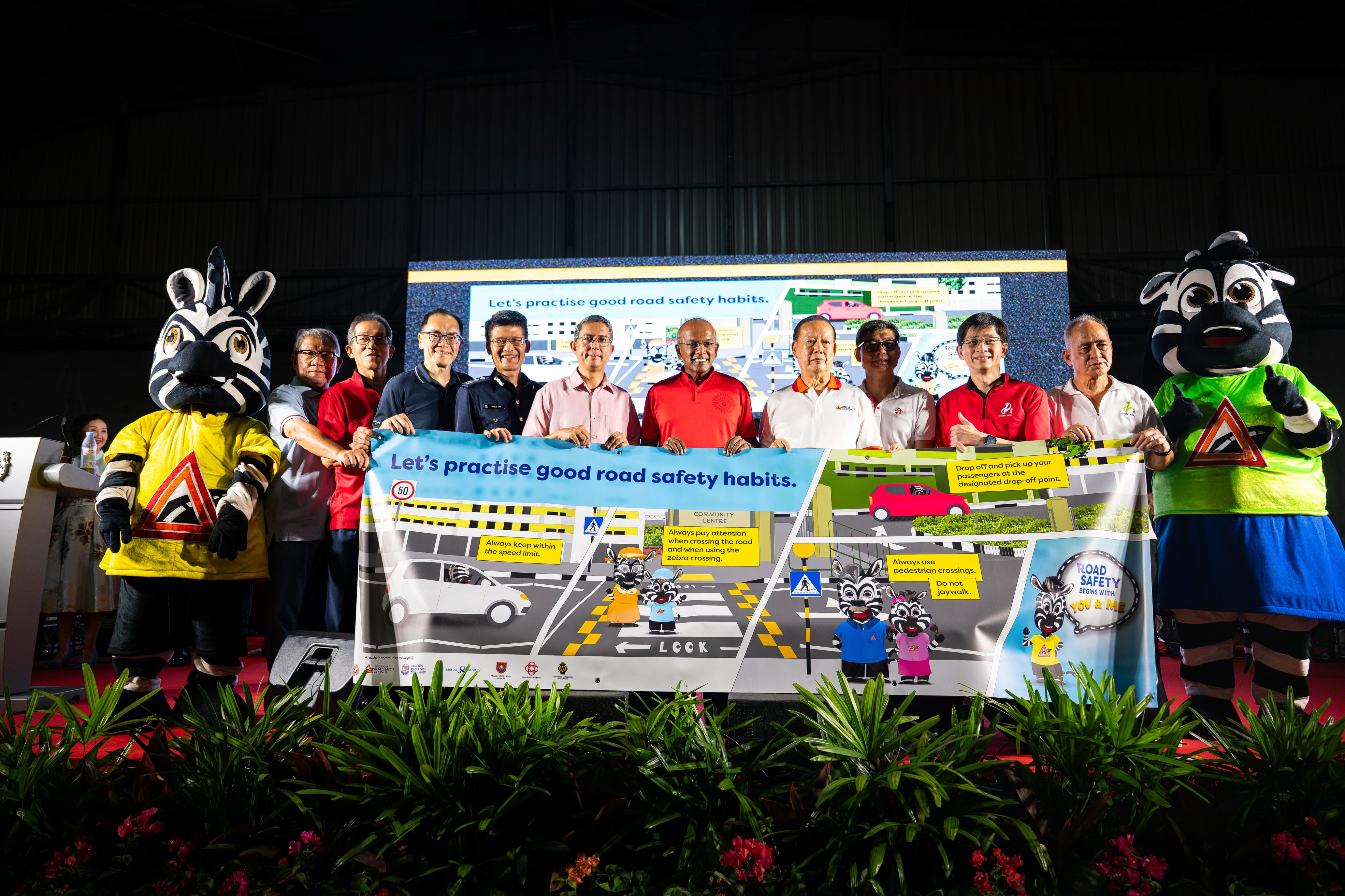 People holding a road safety banner with zebra mascots on each end, in a room with plants in the foreground.