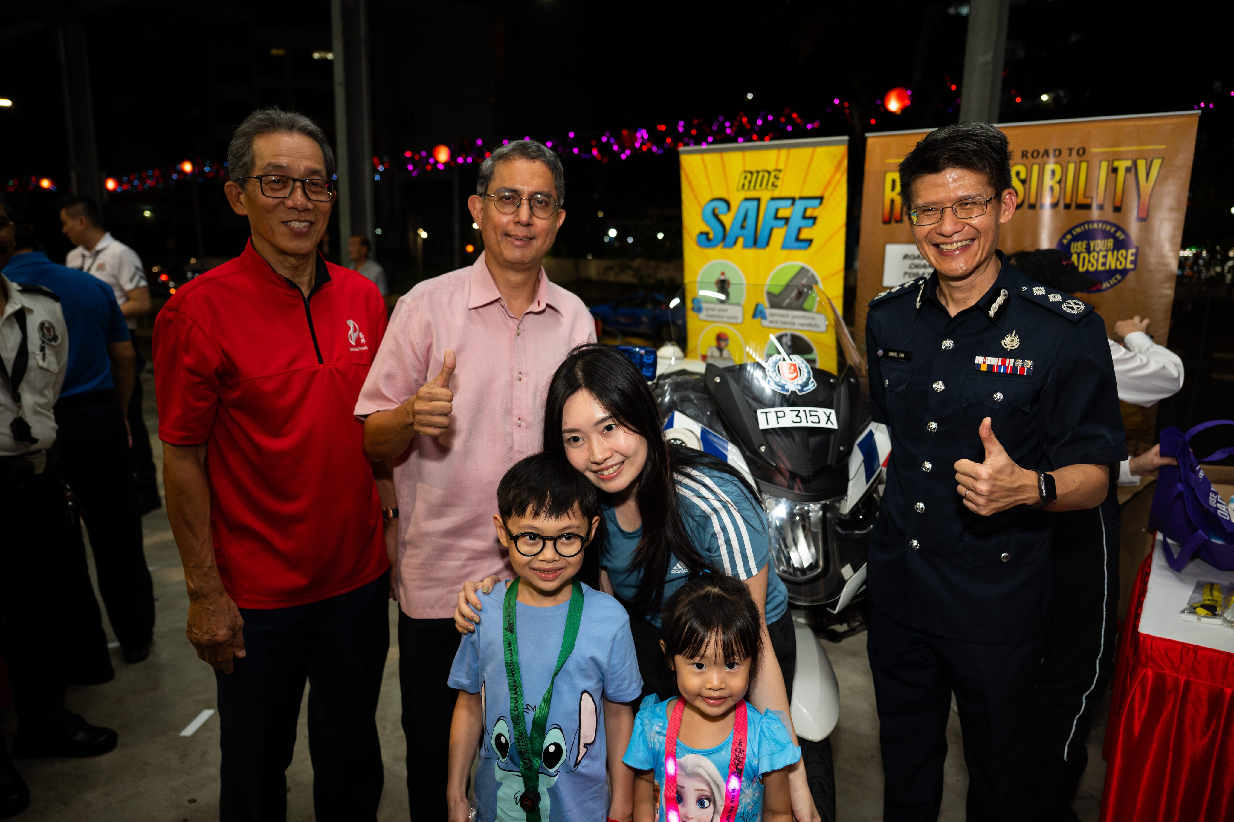 Group of people giving thumbs up, standing in front of a police motorcycle and safety-themed signs.