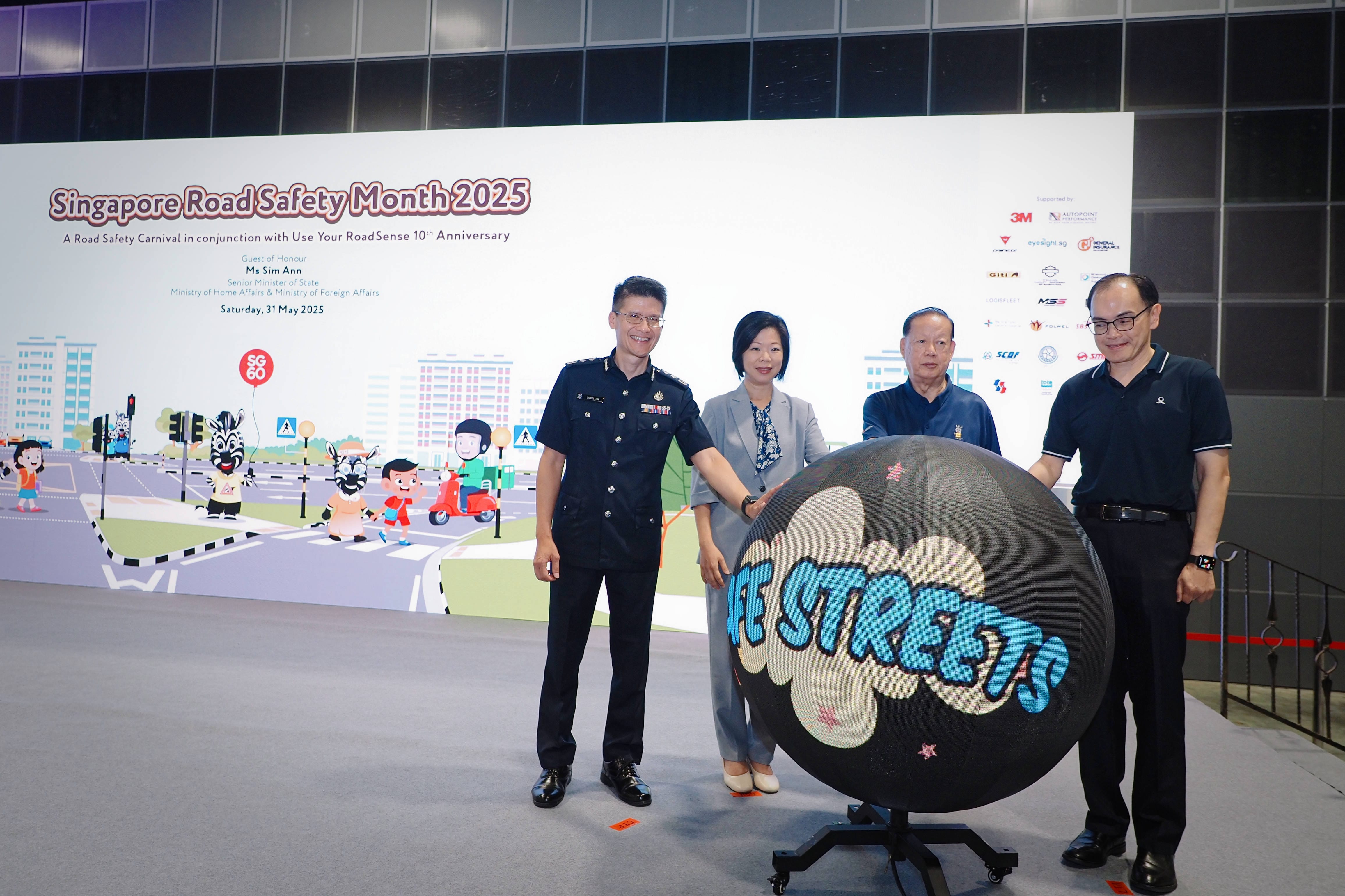 Four people stand in front of a "Singapore Road Safety Month 2025" banner, holding a black sphere with "SAFE STREETS" written on it.