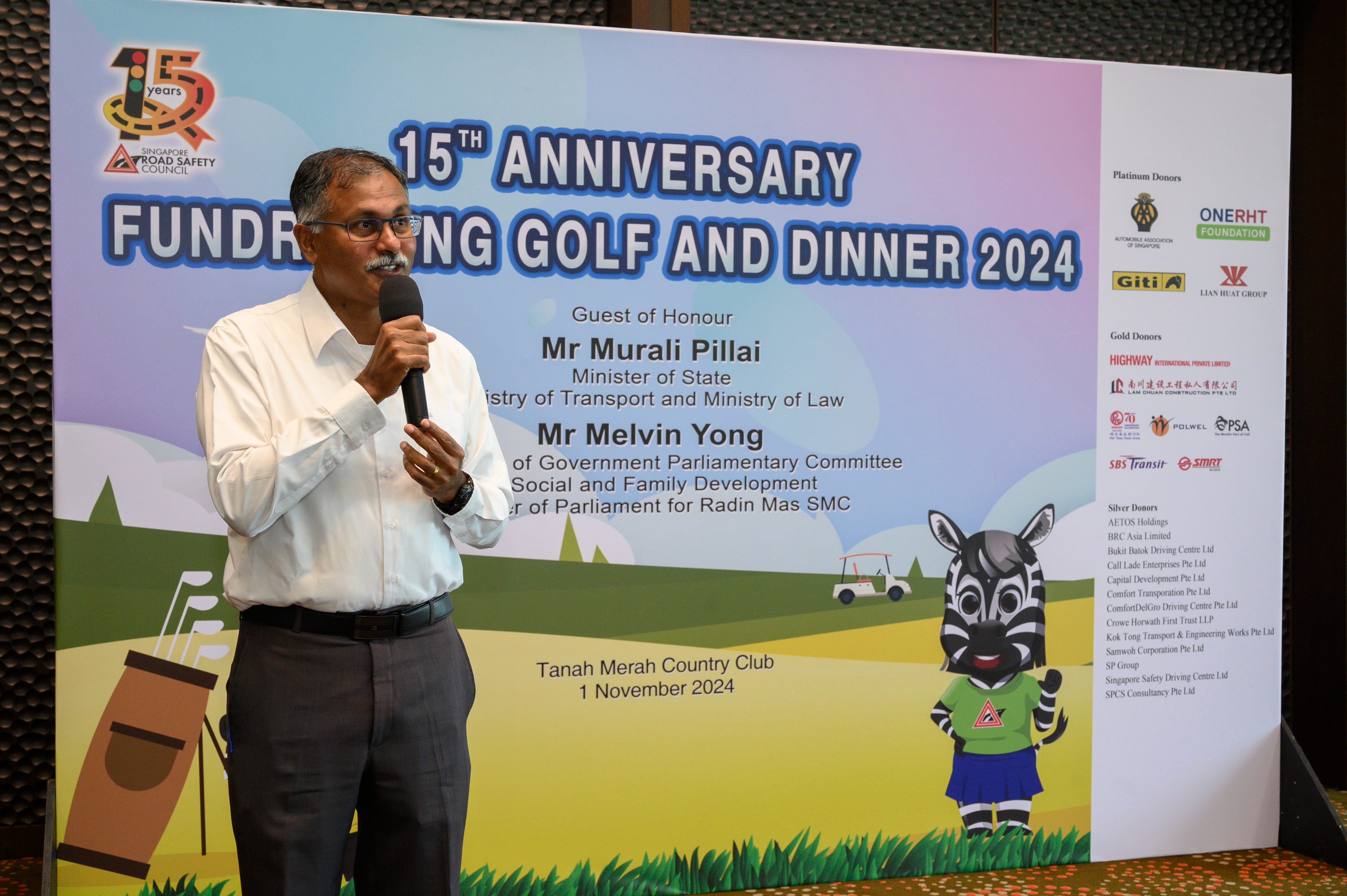 Man in a white shirt speaking into a microphone next to a banner about the 15th anniversary fundraising golf and dinner event.