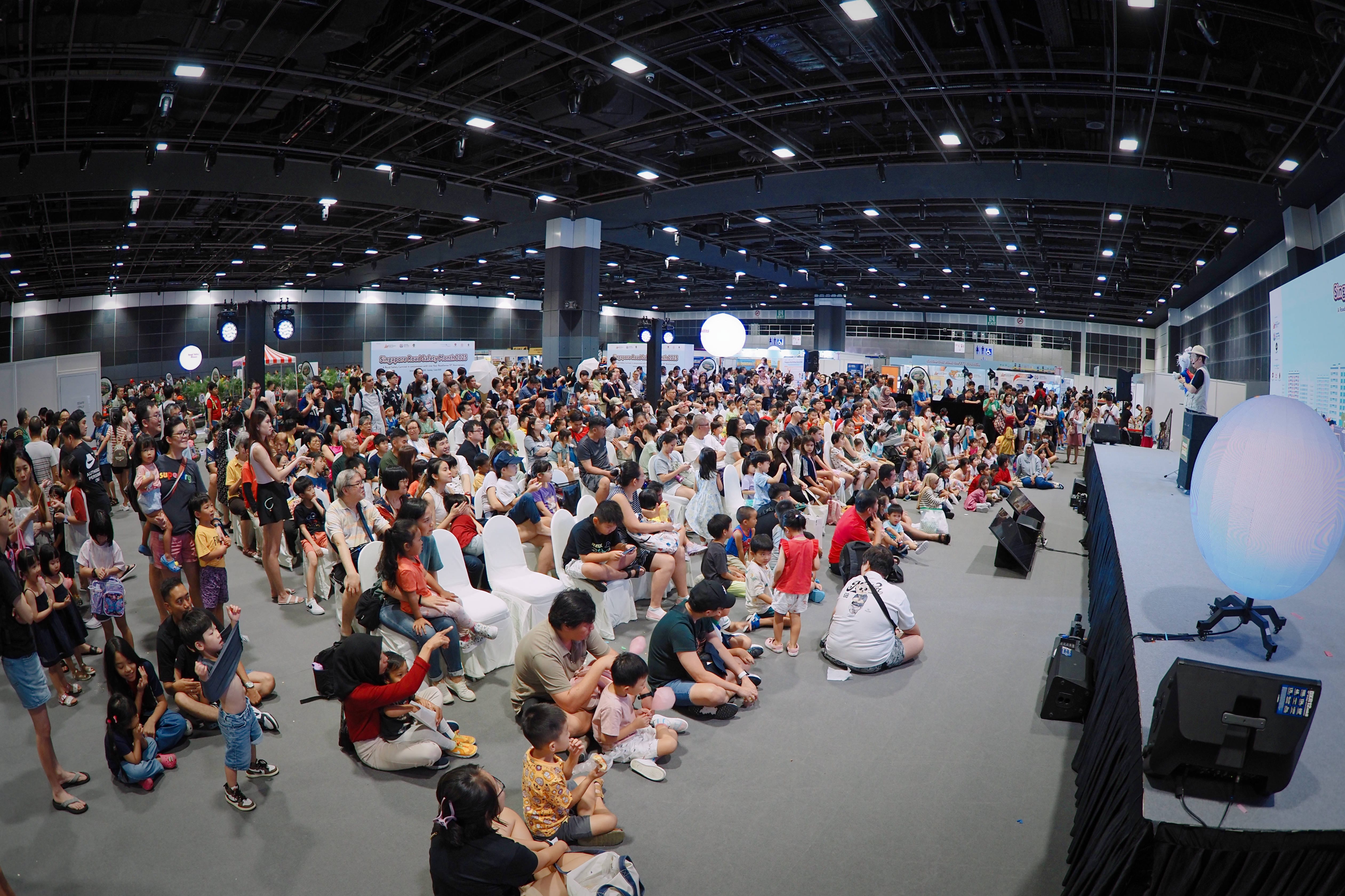 A large crowd of people, including children, sitting and standing in front of a stage indoors, with a speaker on the stage.