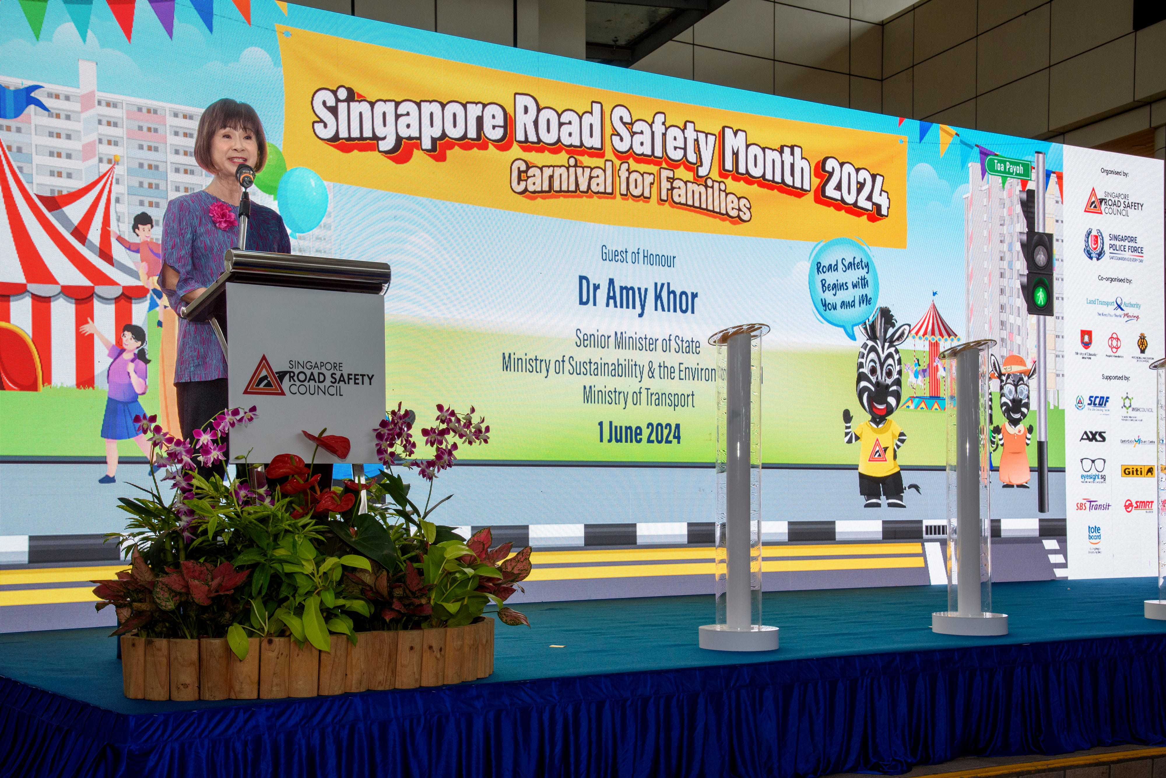 A person is speaking at a podium on a stage with plants, promoting Singapore Road Safety Month 2024, Carnival for Families.