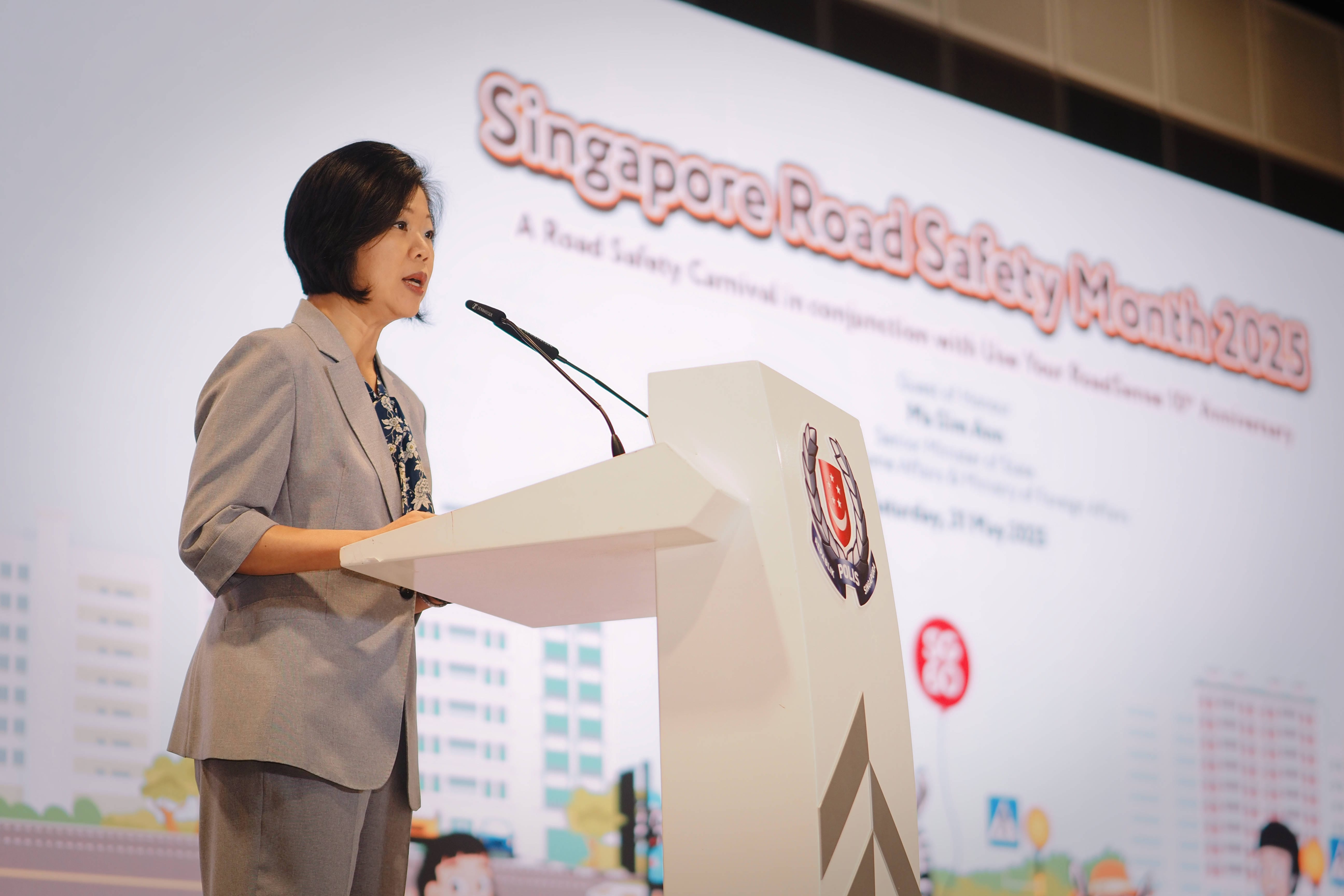 Person in grey suit speaking at podium with Singapore Police Force emblem, backdrop reads "Singapore Road Safety Month 2025".
