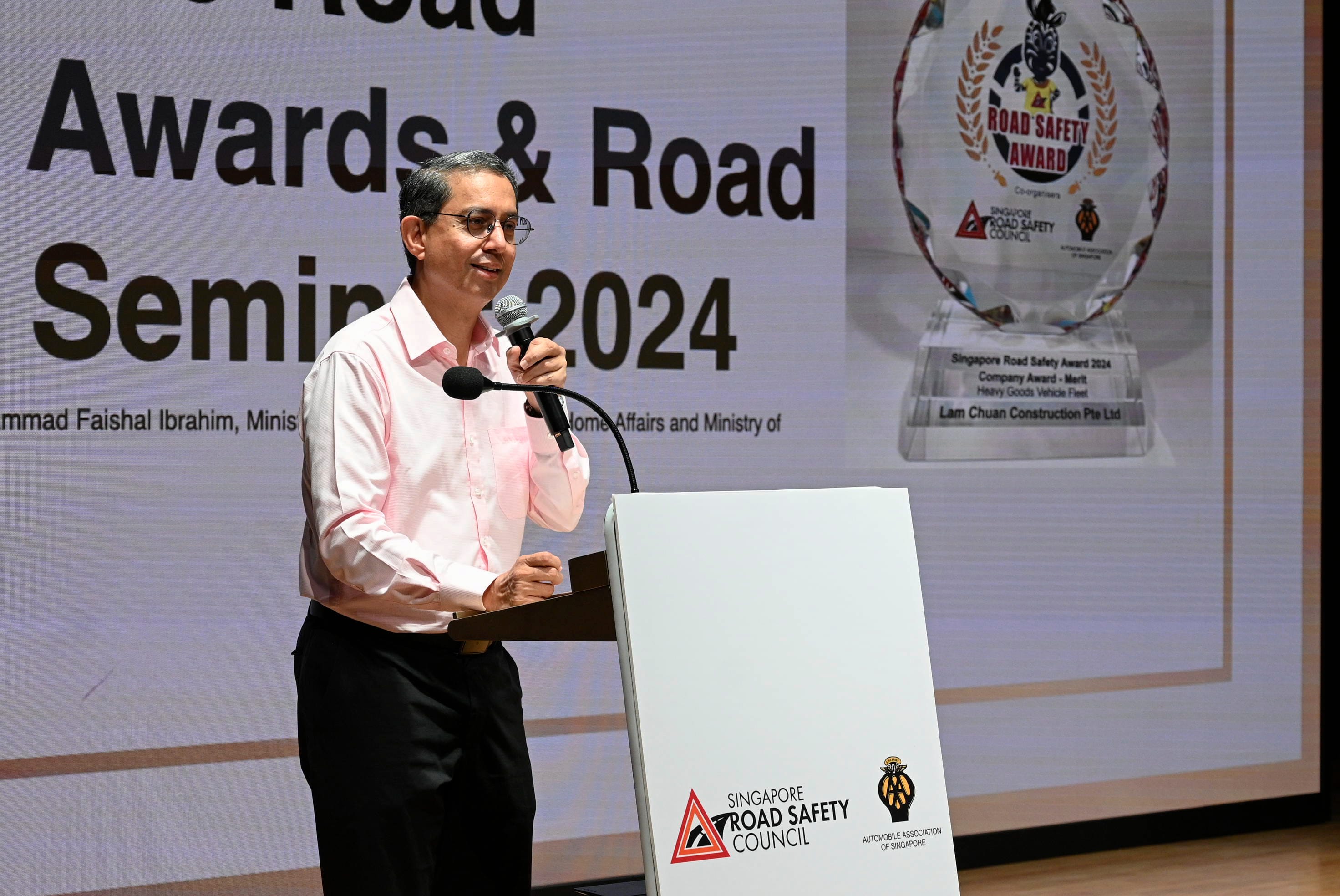 A person speaking at a podium with a microphone, with a Singapore Road Safety Council banner and an awards display behind.
