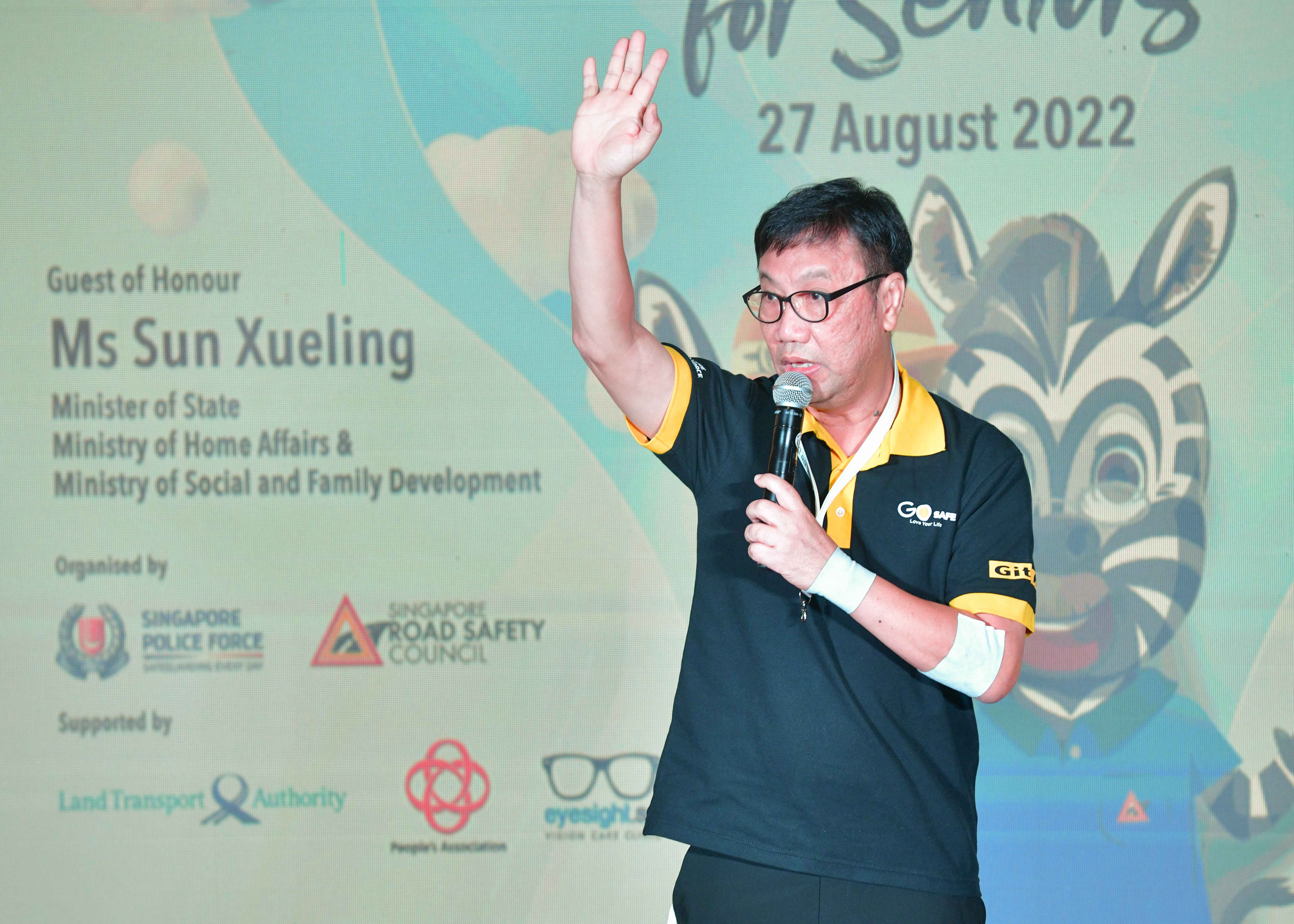 A person in a black shirt holding a microphone and raising their hand, standing in front of a presentation backdrop.