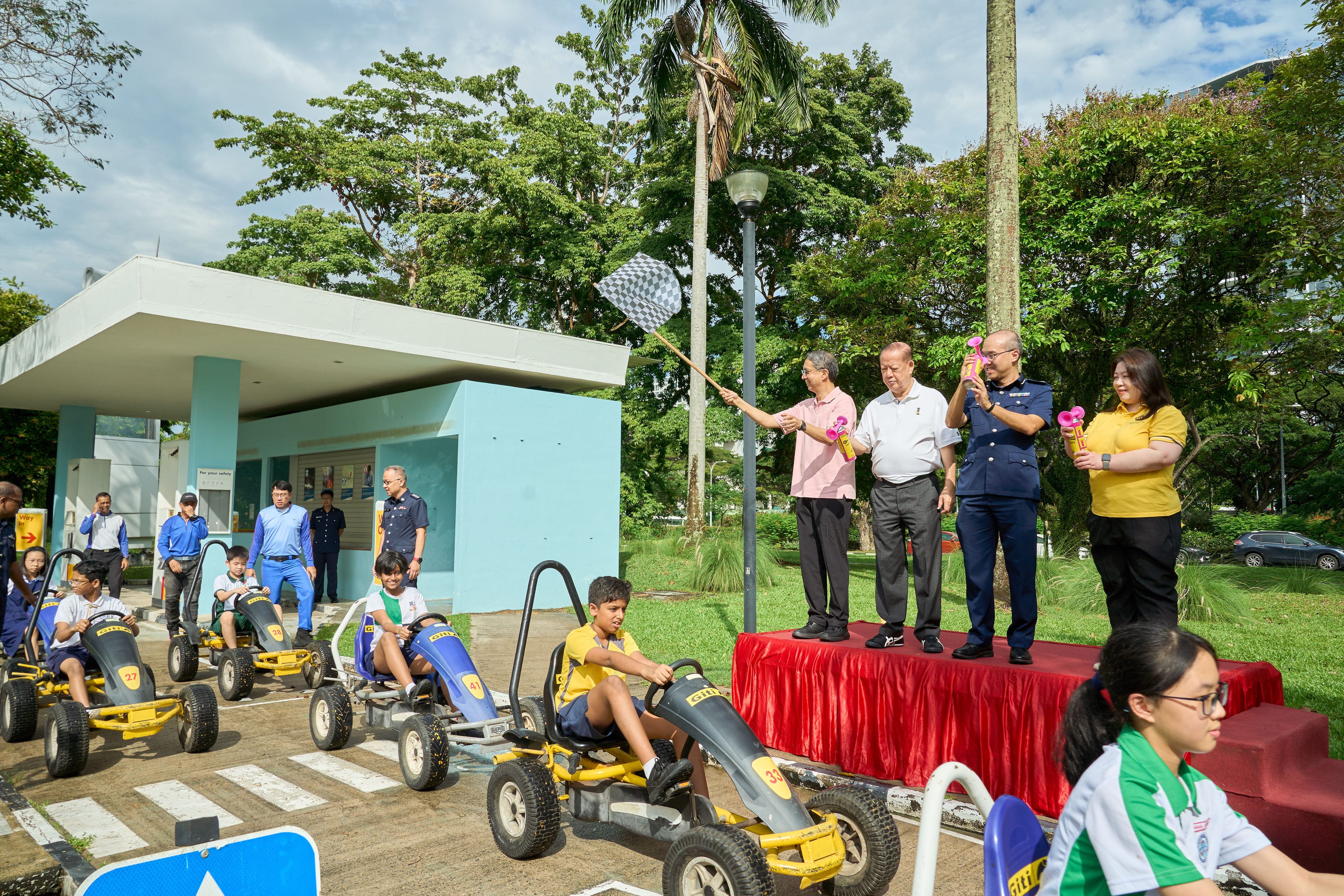 People in blue and yellow shirts stand near four pedal karts at a race starting line, outdoors on a sunny day.
