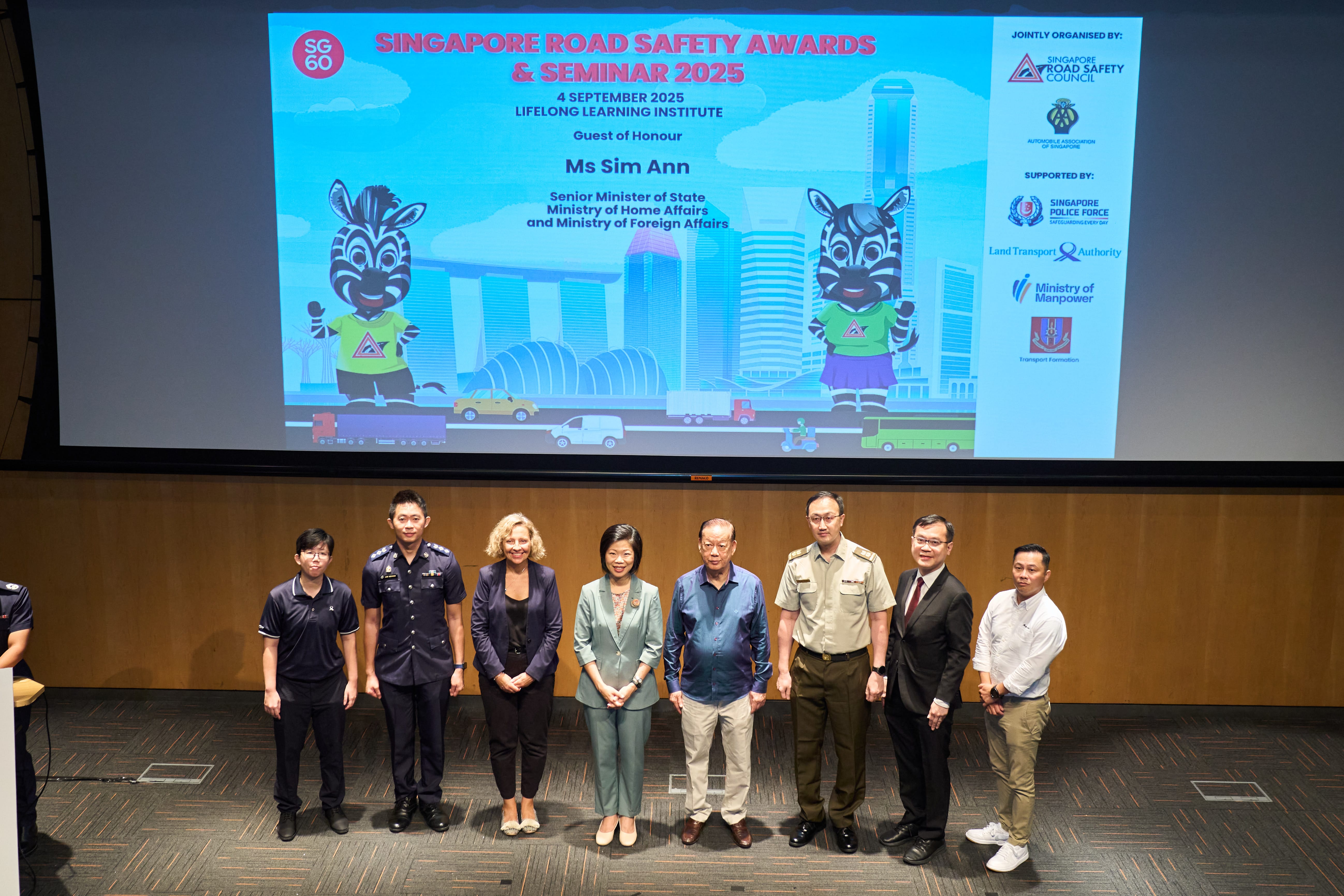 Eight people in formal attire standing on a stage with a large backdrop promoting the Singapore Road Safety Awards and Seminar 2025.