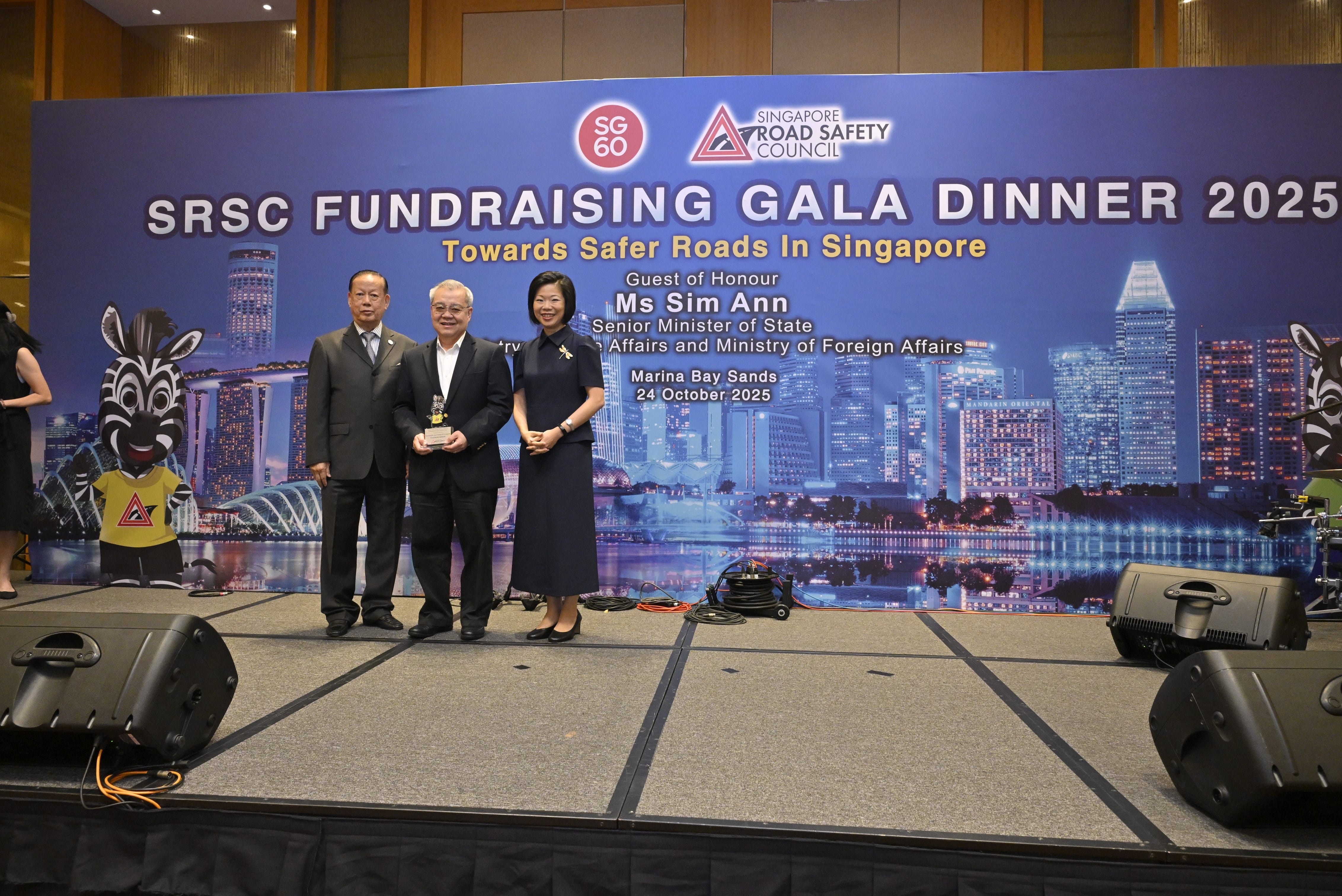 Three individuals stand on a stage, with a zebra mascot and city skyline background promoting Singapore Road Safety Council.