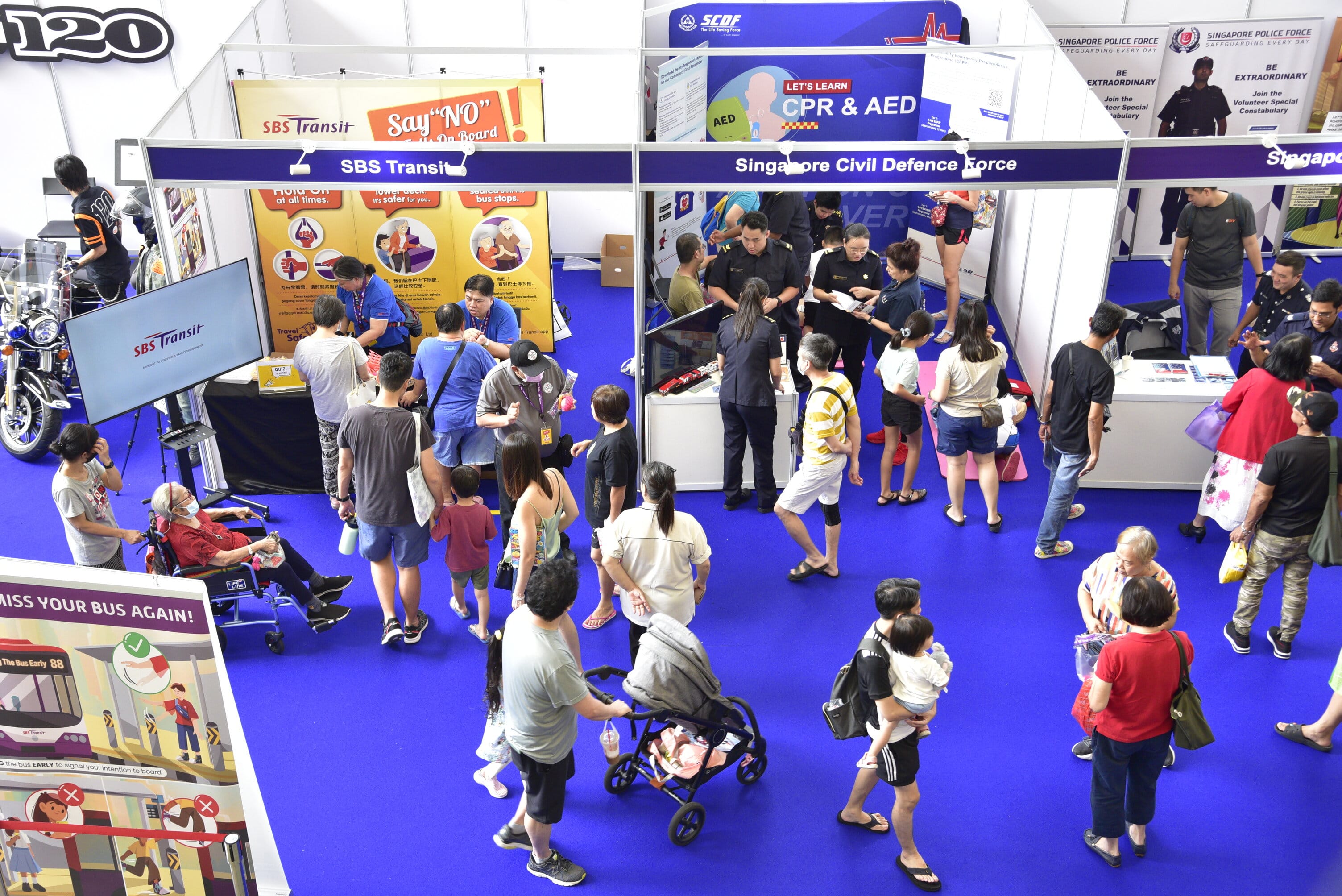 A crowd visits various informational booths on safety, CPR, and police recruitment at an indoor event with a bright blue carpet.