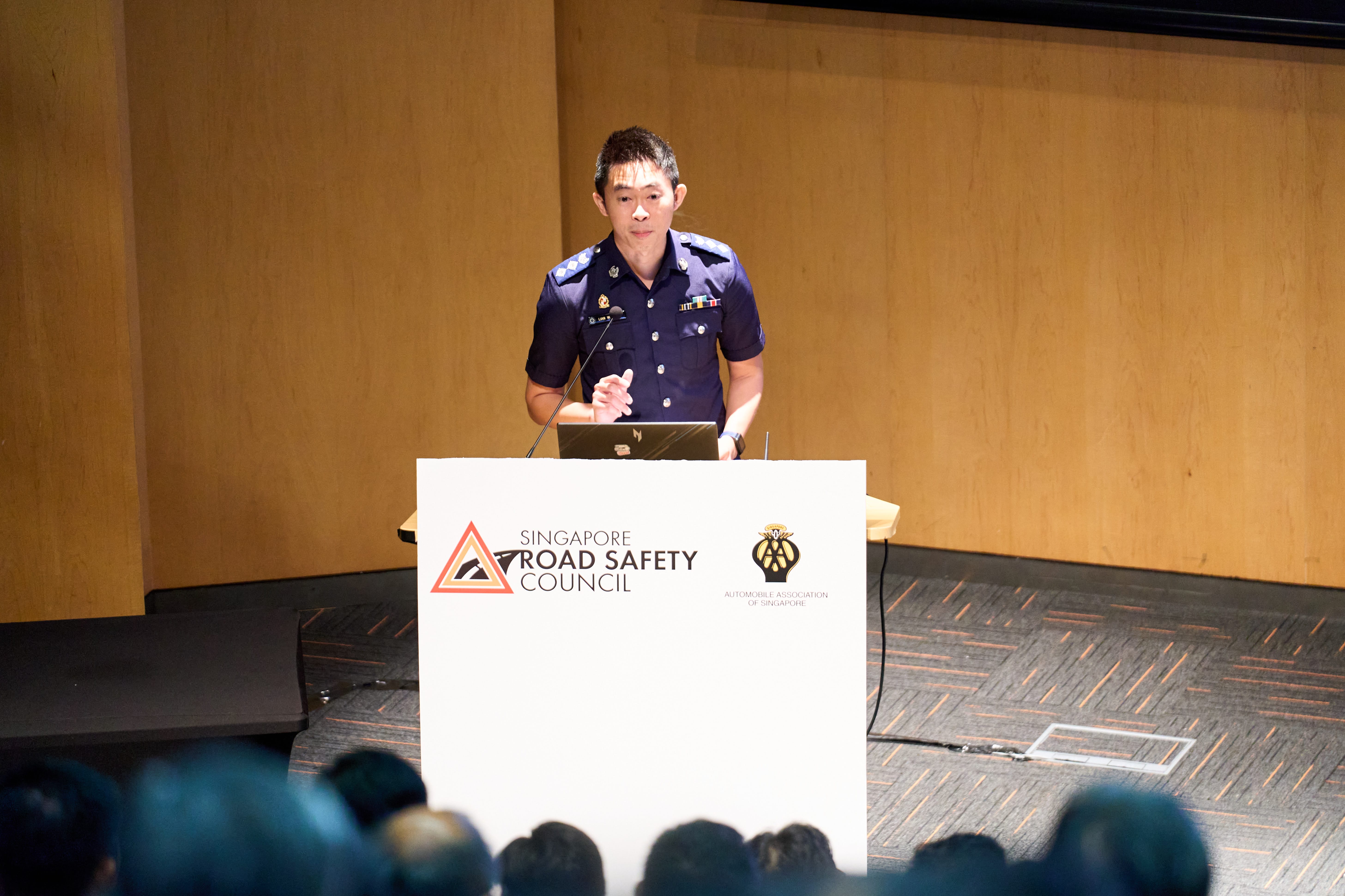 A uniformed person standing at a podium with a "Singapore Road Safety Council" sign, addressing an audience.