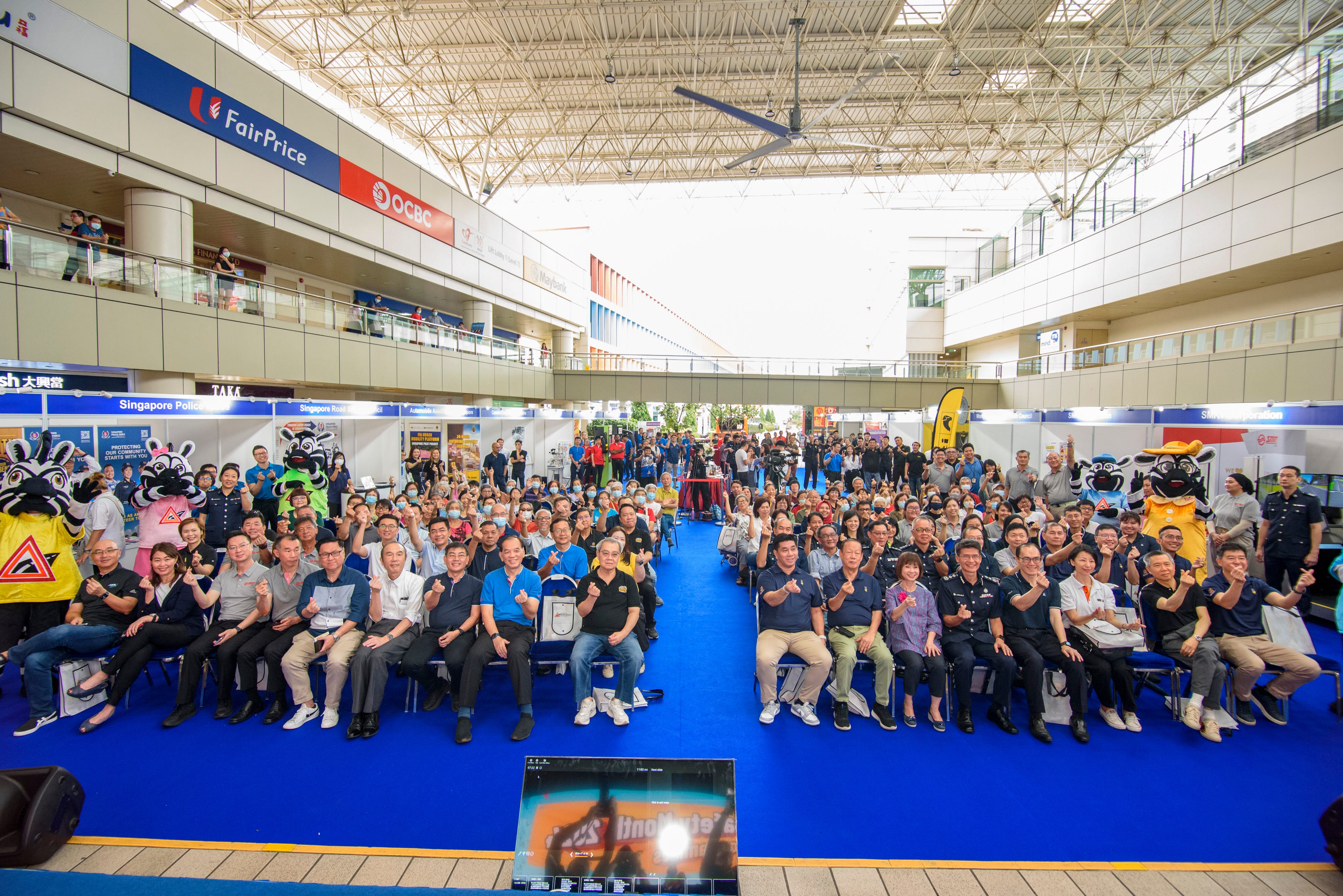 A large group of people sitting and standing on a blue carpet in an indoor event space, with various booths and mascots present.