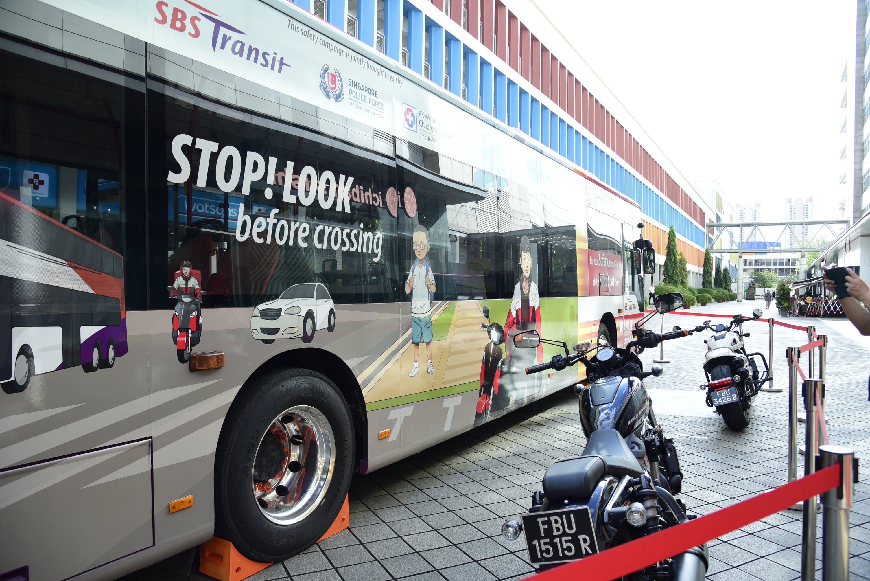 A bus with safety campaign graphics is parked near motorcycles, with the text "STOP! LOOK before crossing" on its side.