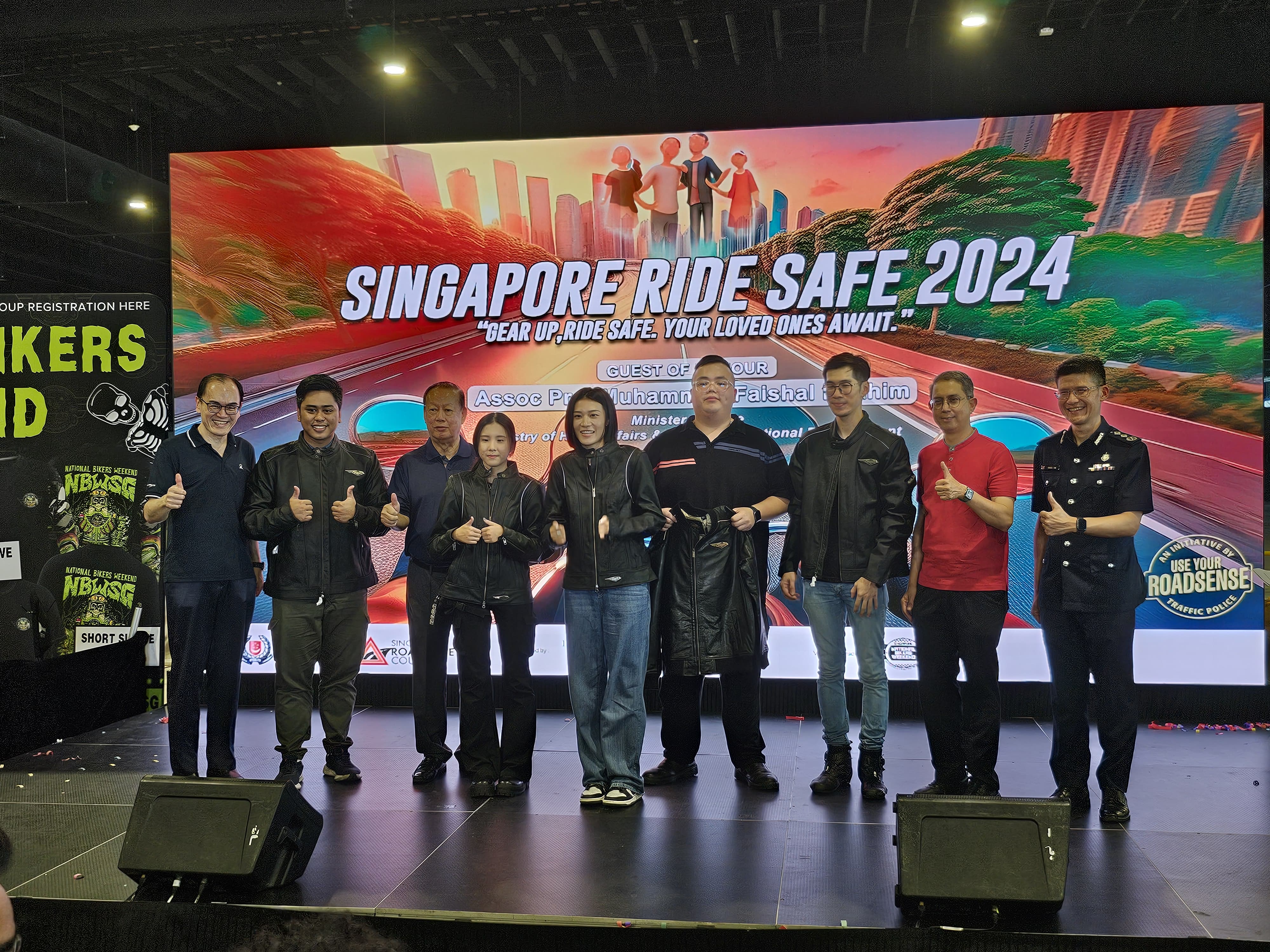 A group of people on stage giving thumbs up in front of a "Singapore Ride Safe 2024" event banner.