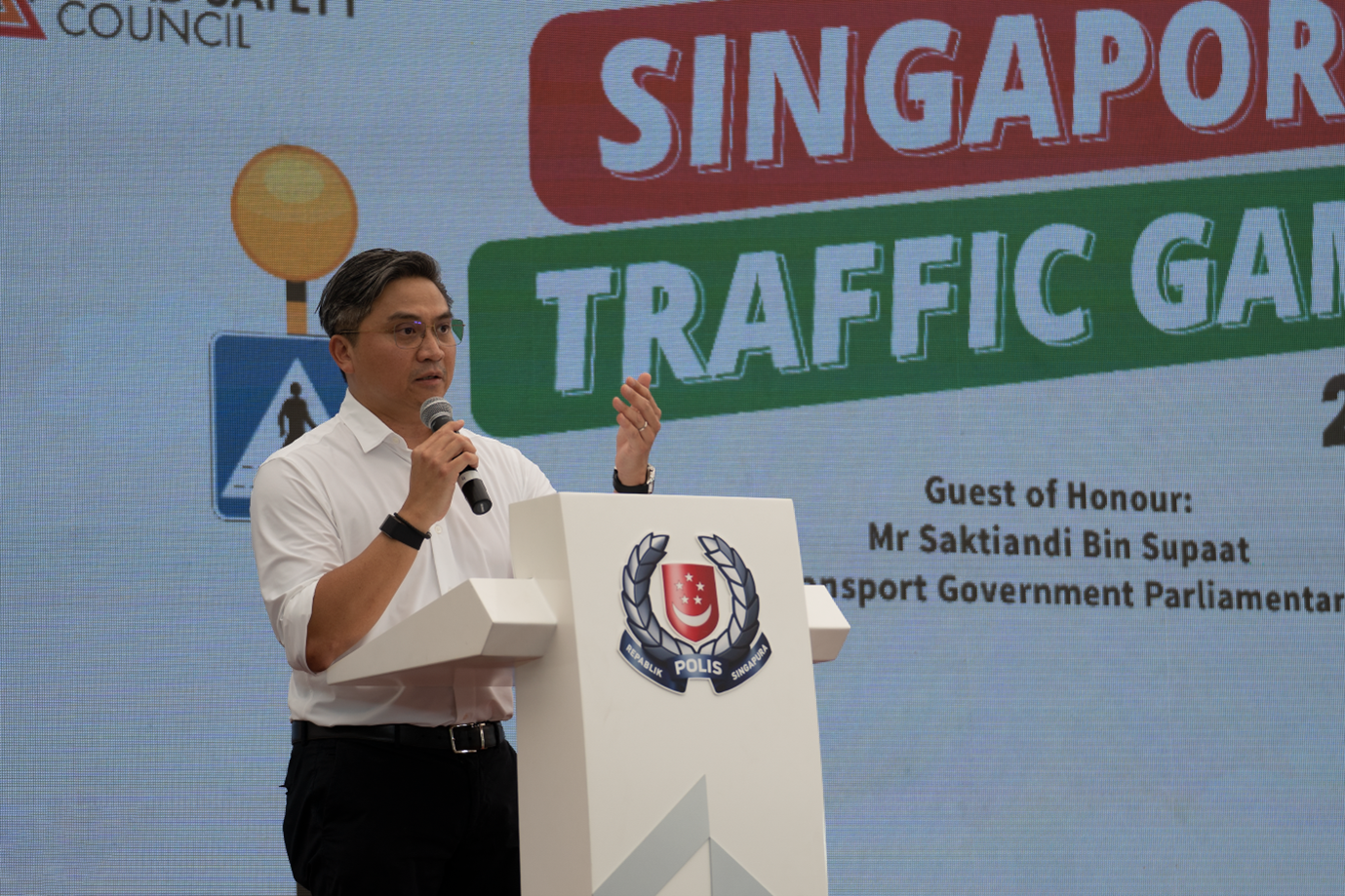 A person in a white shirt is speaking at a podium with a Singapore Police emblem, beside a large "Singapore Traffic Games" sign.