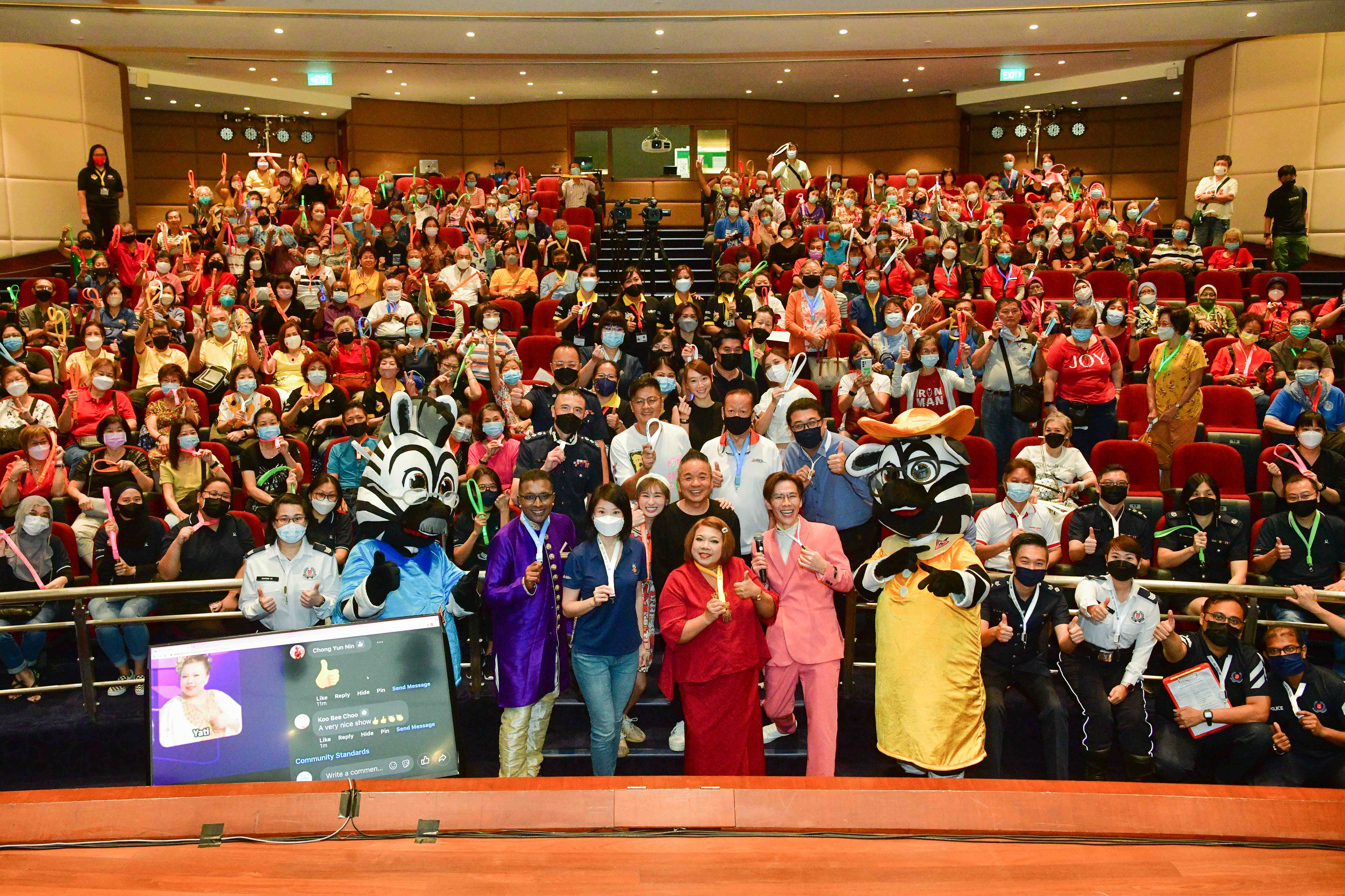 A large group of people in an auditorium with colorful outfits, some in costume, holding various light sticks and objects.