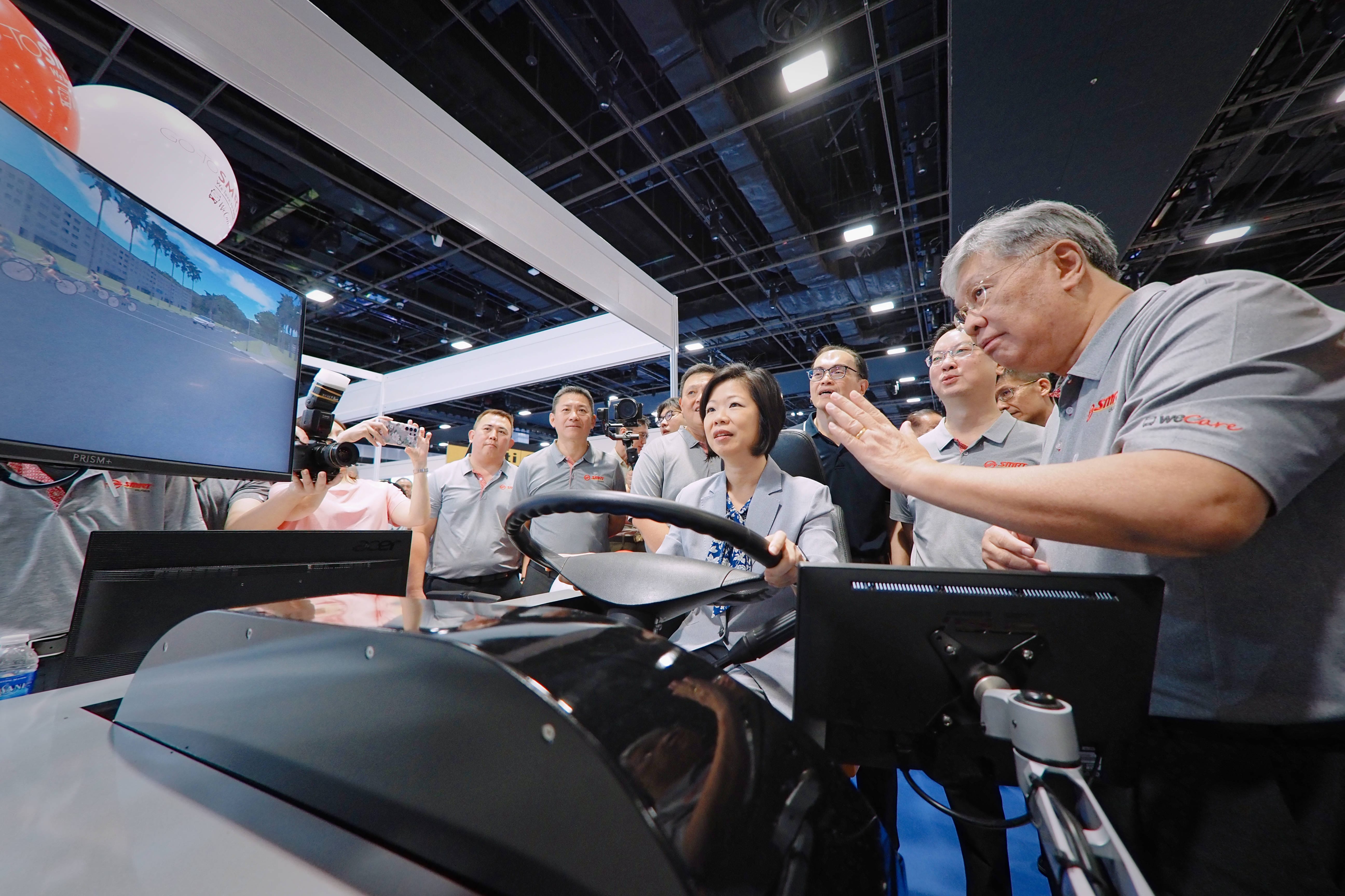 A group of people gathers around a driving simulator with multiple screens at a tech exhibit.