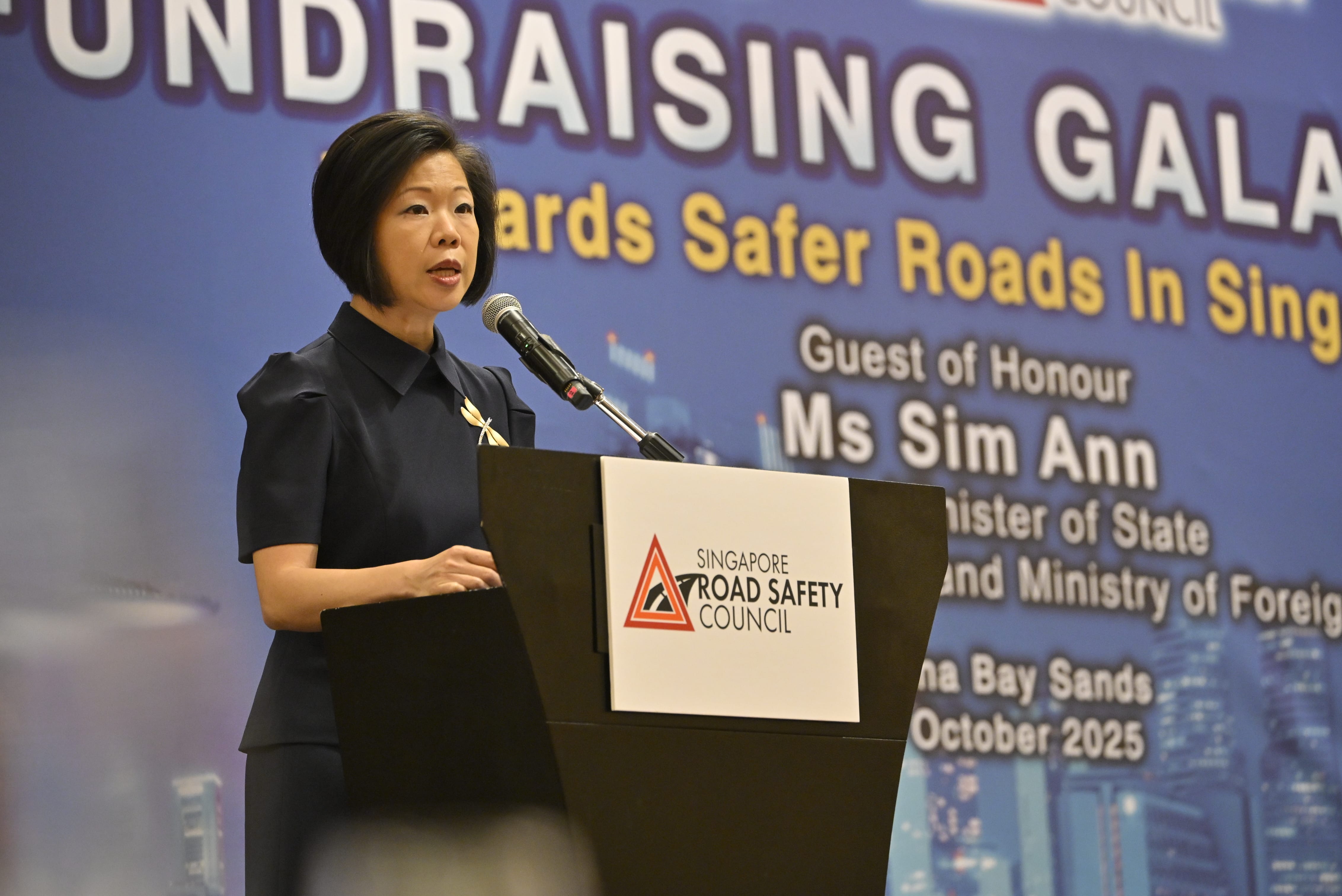 A person is speaking at a podium with a sign for the Singapore Road Safety Council, against a backdrop showing event details.