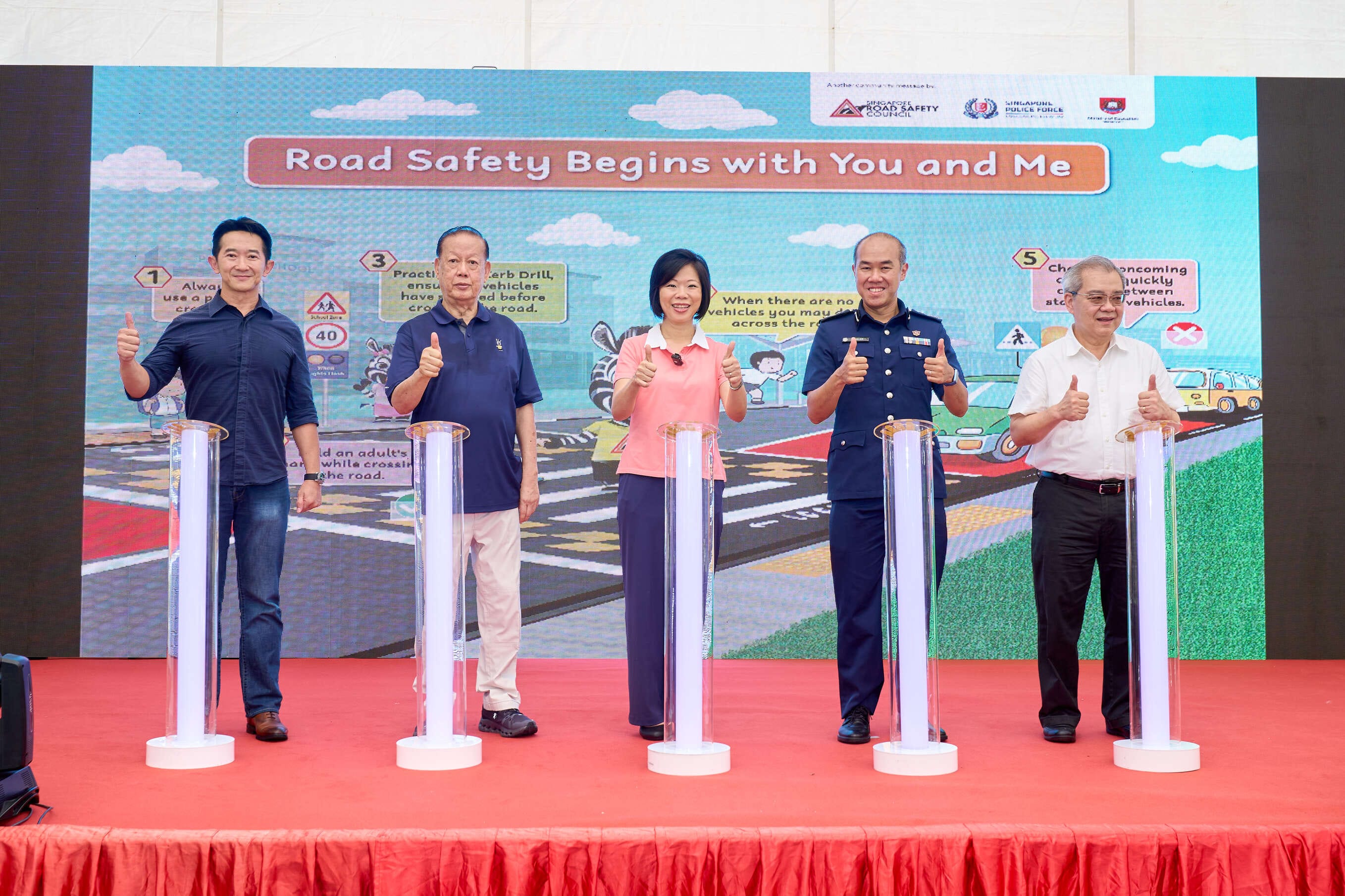 Five people give thumbs up next to lighted pillars on a stage with a road safety backdrop.