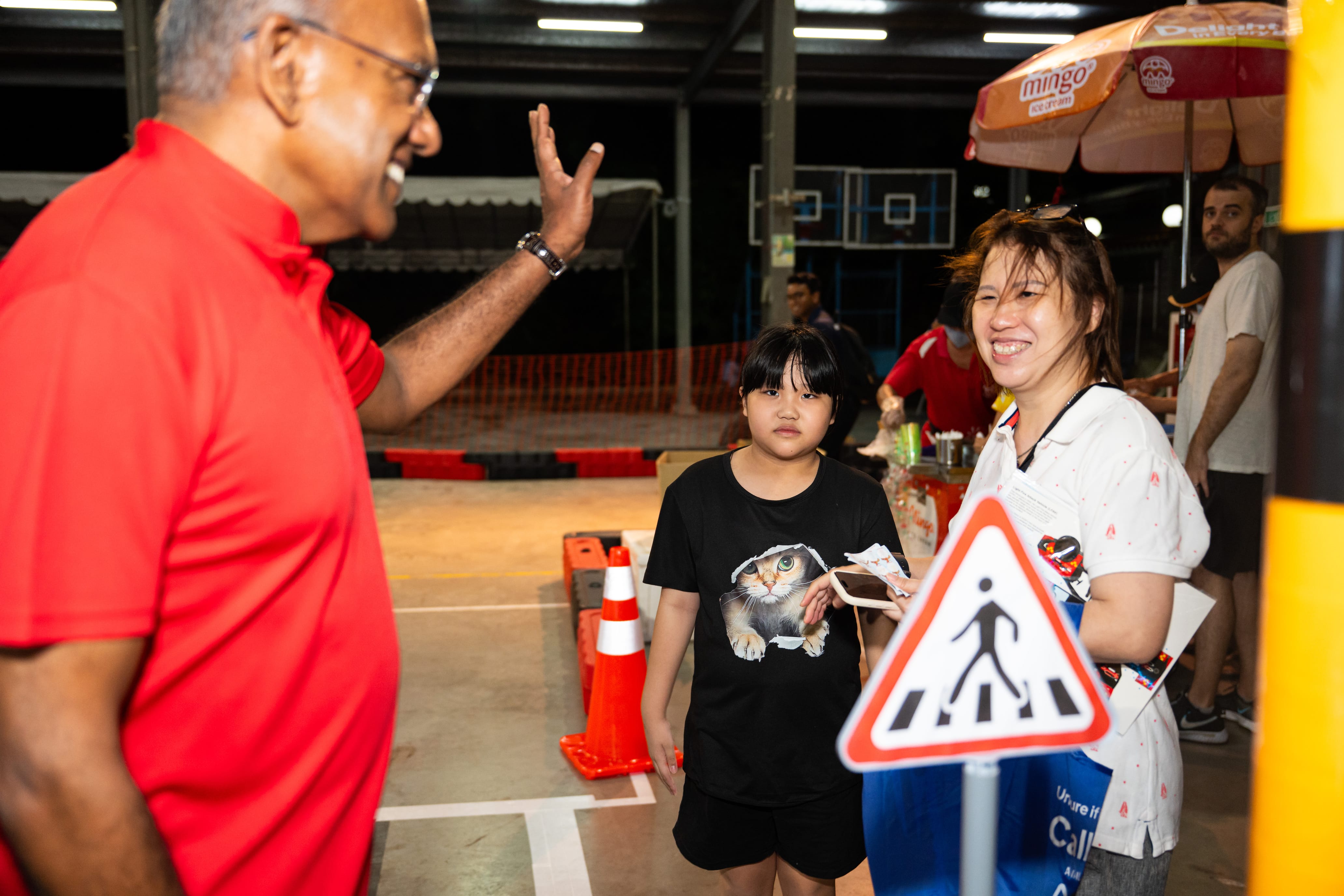 A person in a red shirt gestures near a walking sign with a cat print shirt and another person holding items under an umbrella.
