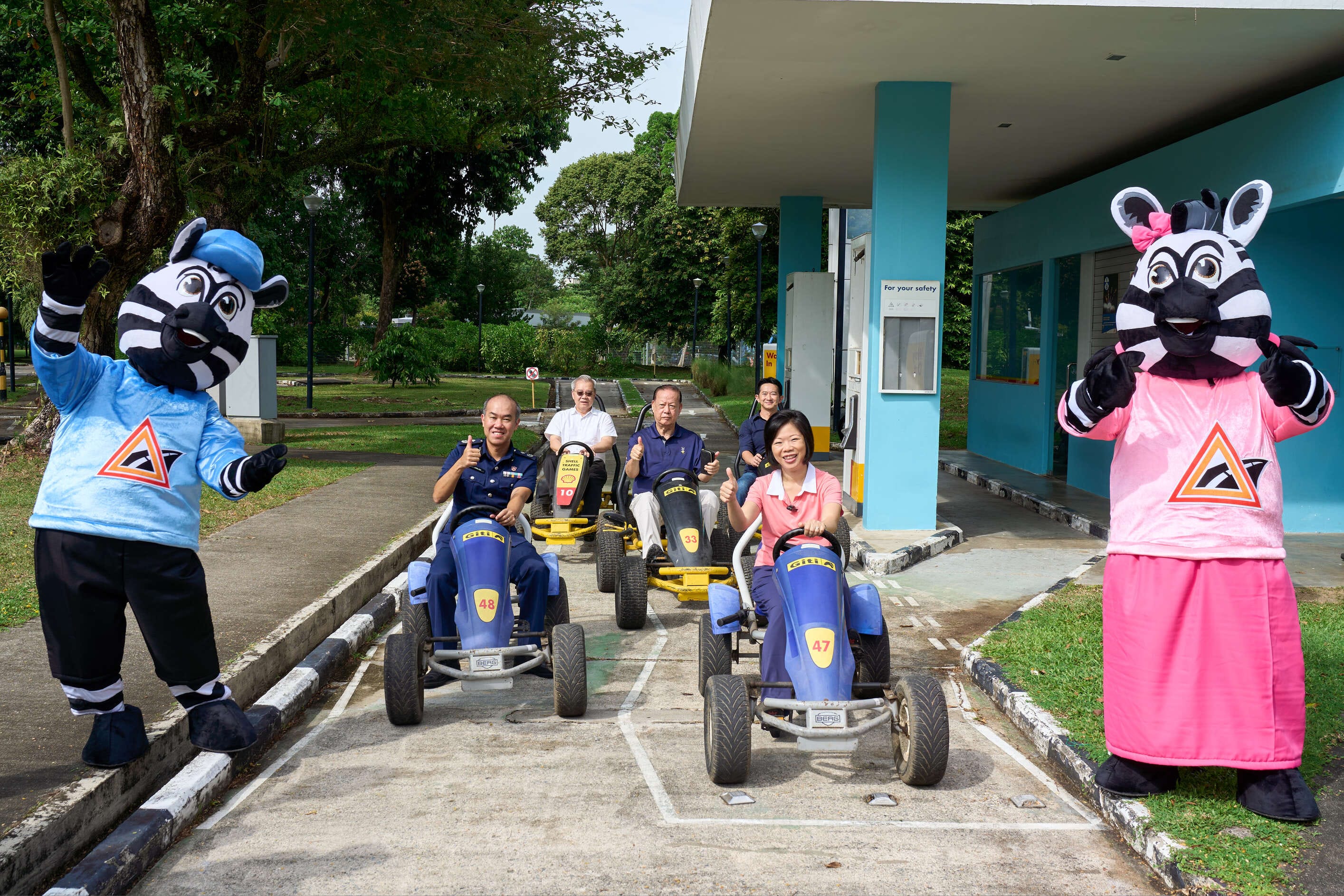 Two people in zebra costumes stand beside a group riding go-karts at a park with trees and a building in the background.