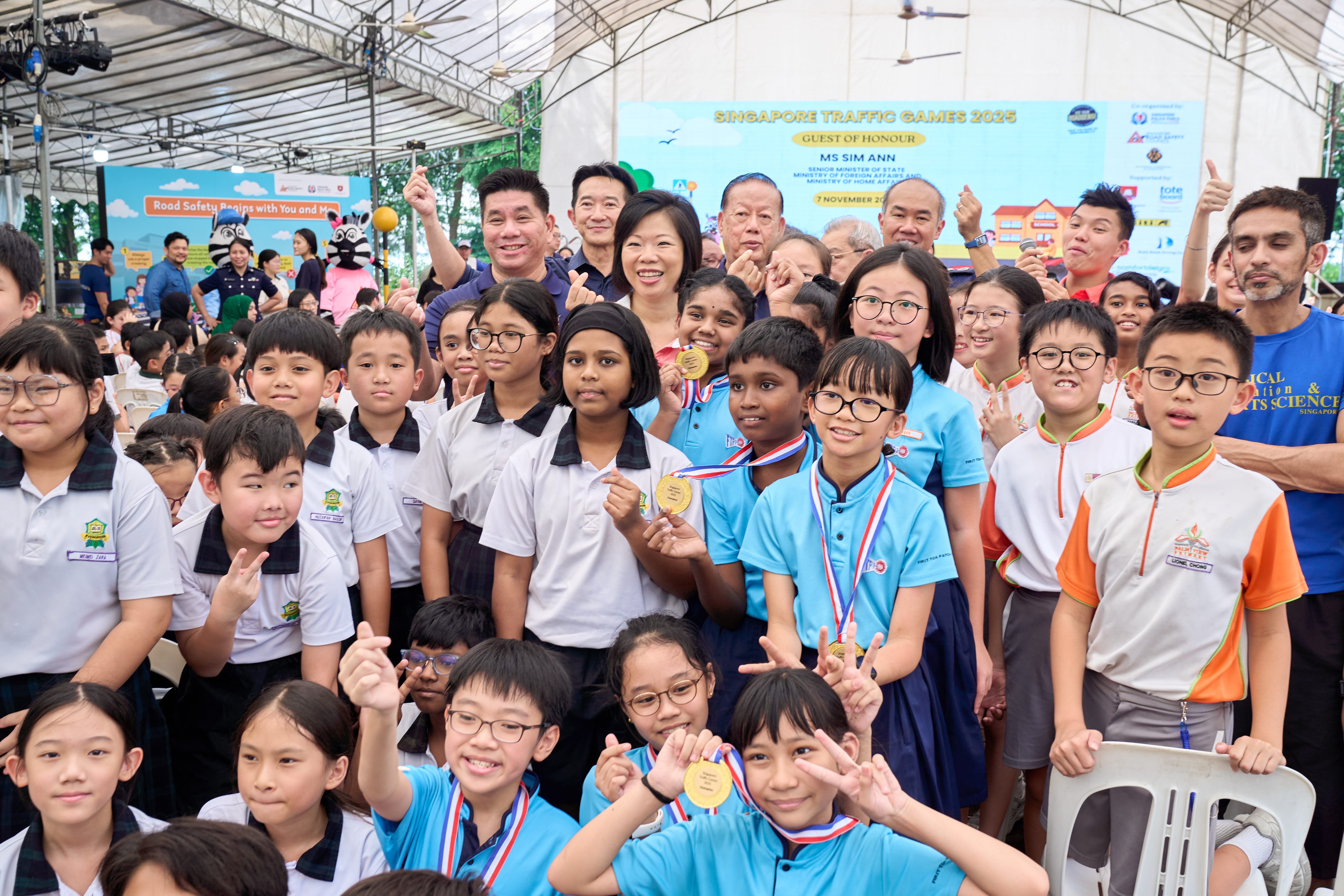 A group of students wearing uniforms gather indoors, holding medals; a banner in the background reads "Singapore Traffic Games 2023."