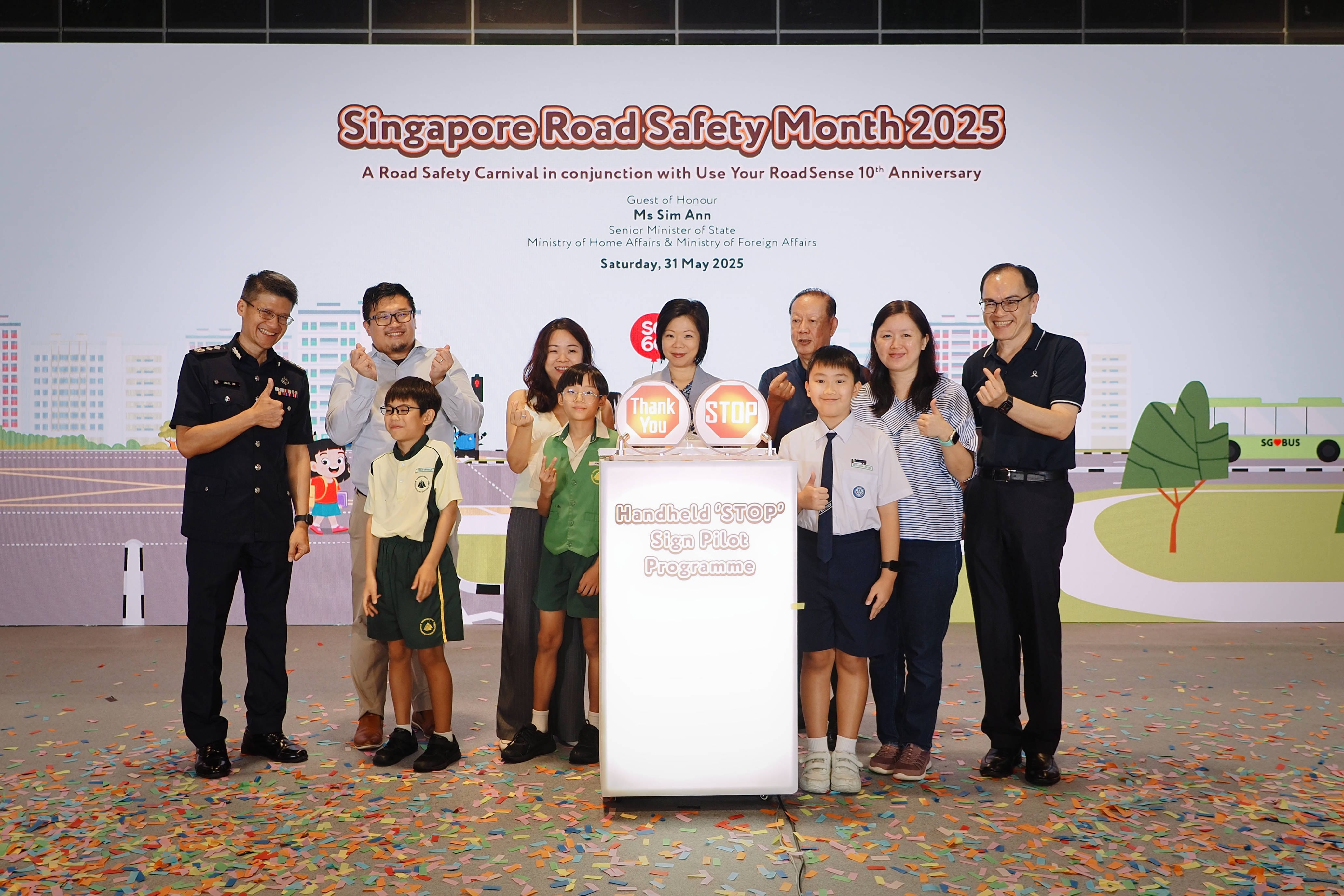A group of people stands in front of a "Singapore Road Safety Month 2025" backdrop with confetti on the ground.