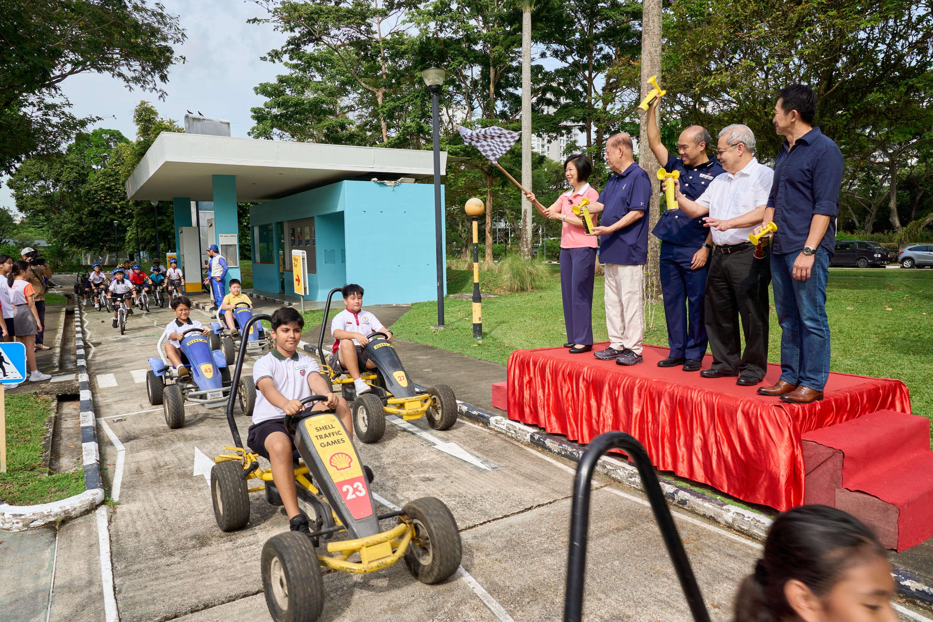 A group of people holding flags and trophies stand on a platform while children participate in go-karting on a track.
