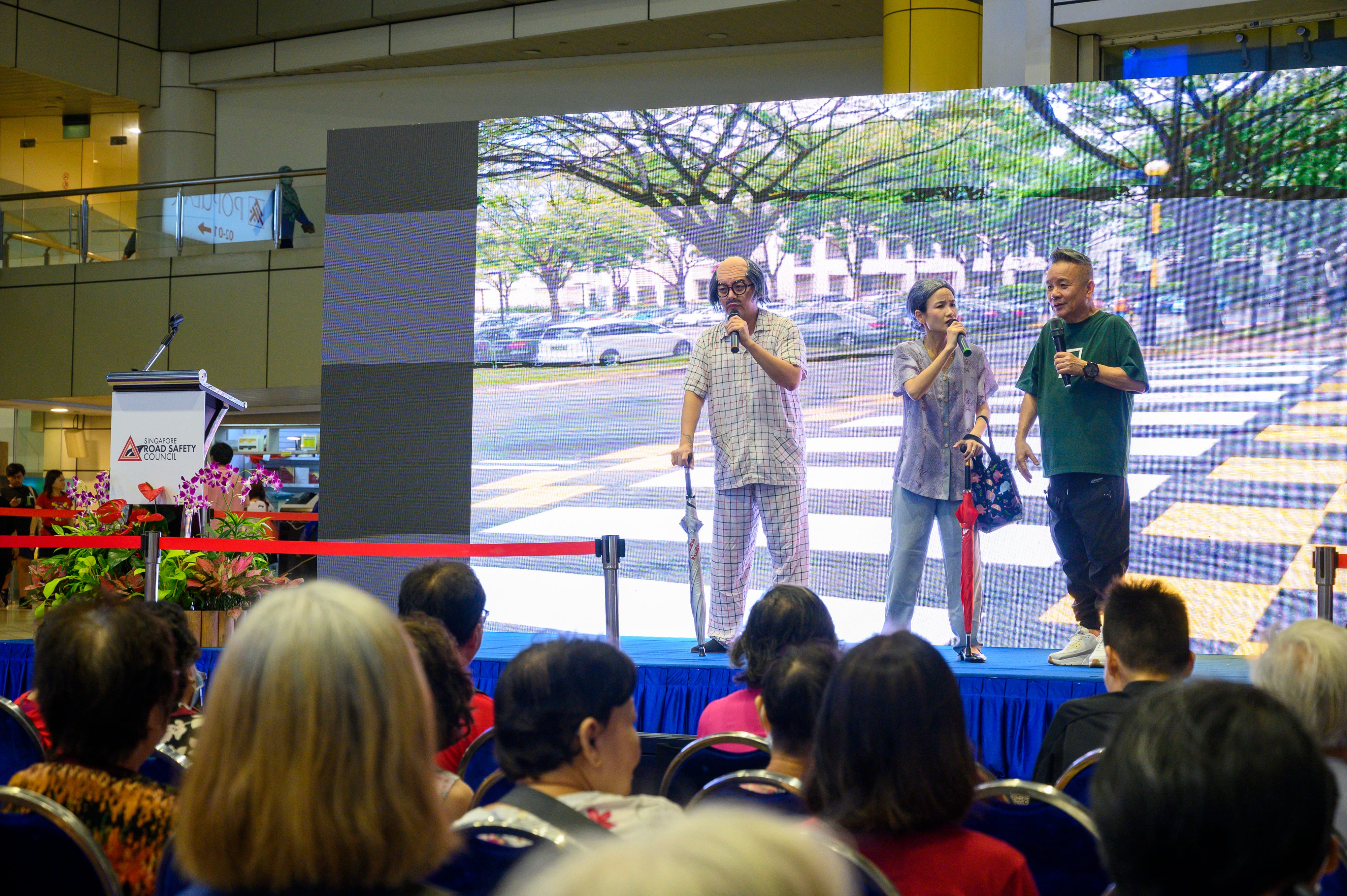 Three people stand on a stage with a large screen backdrop and an audience seated in front.
