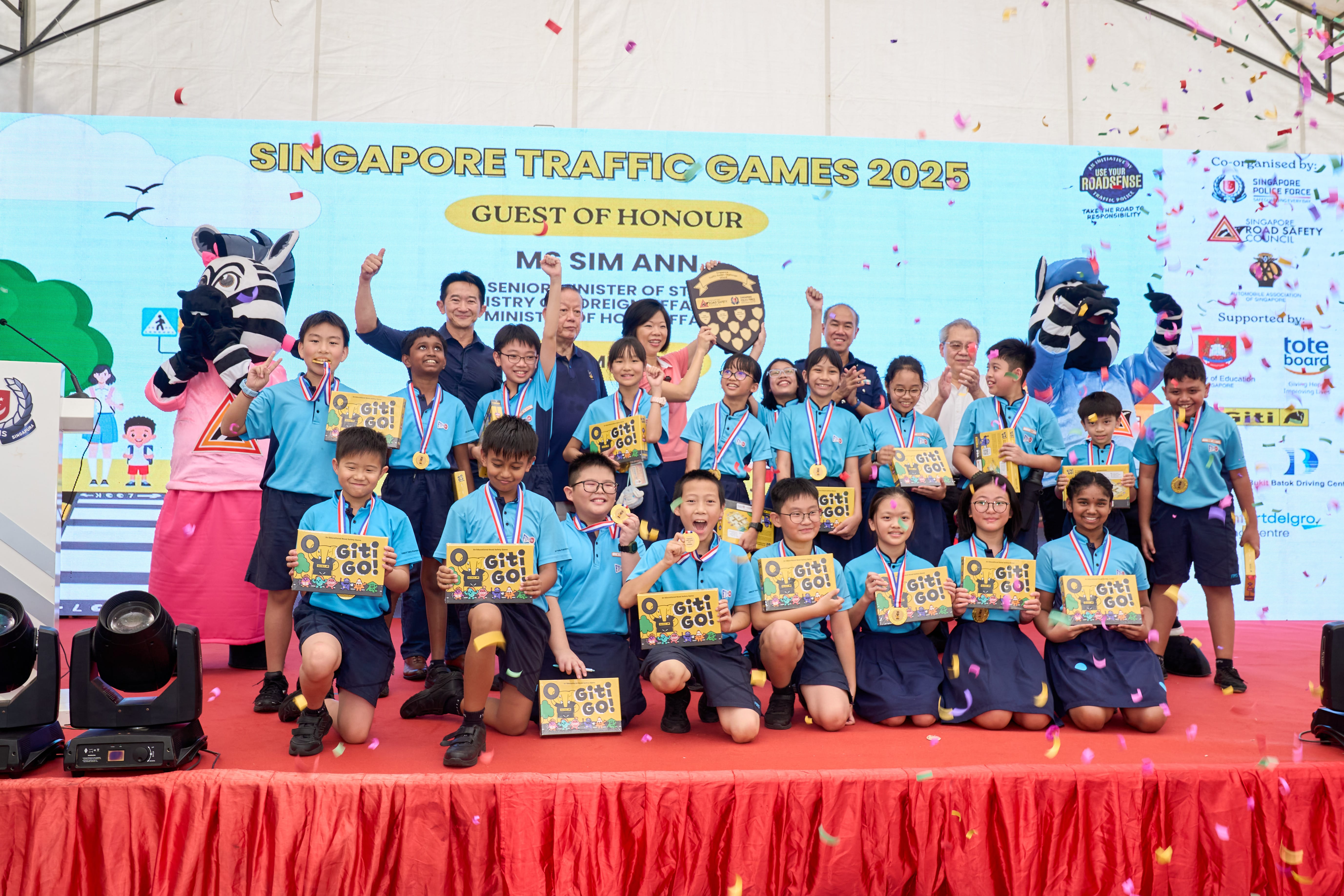 Children in blue uniforms stand on a stage holding certificates, surrounded by colorful confetti with two mascots nearby.