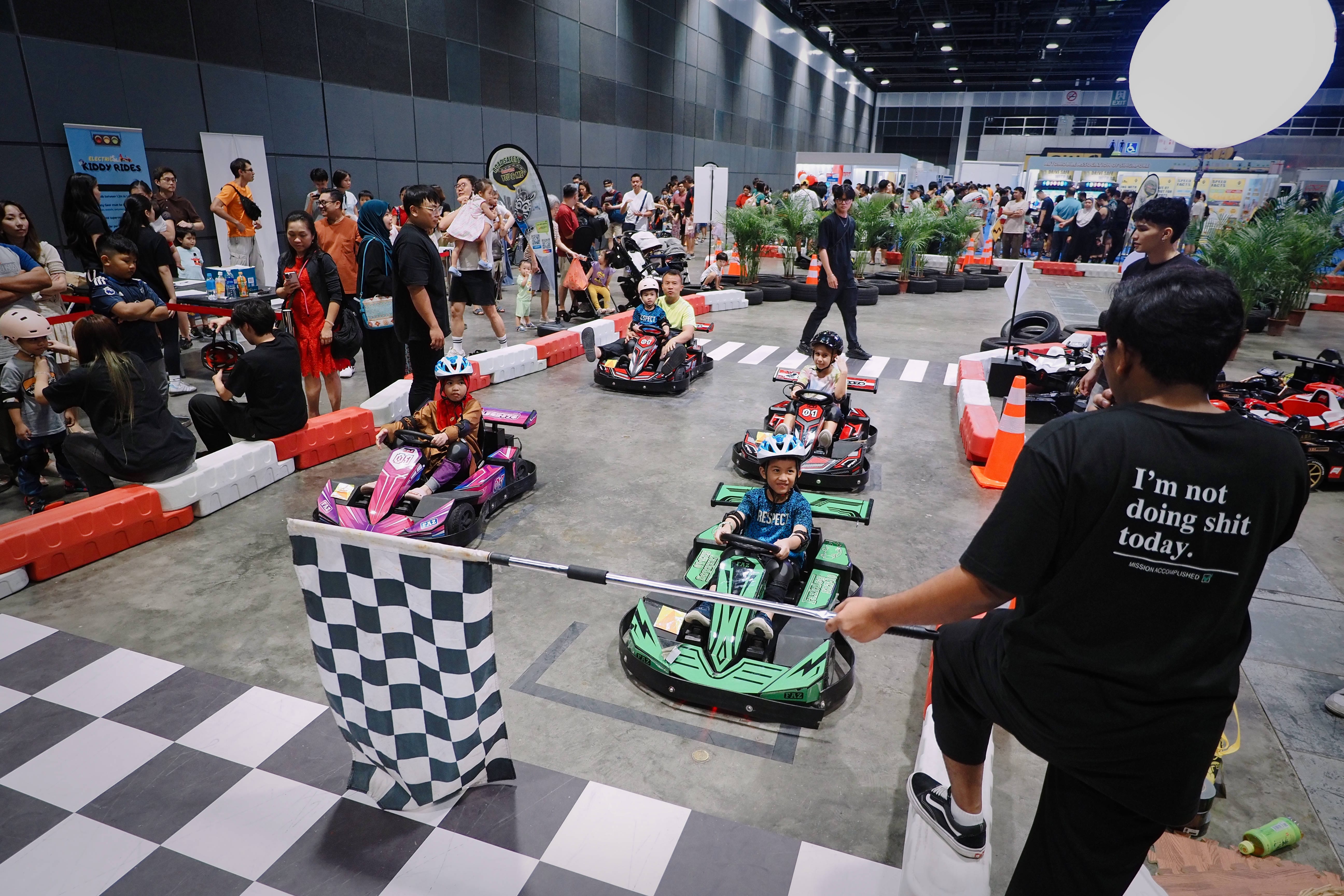 Children in miniature go-karts lined up for a race, surrounded by spectators and barriers in an indoor venue.