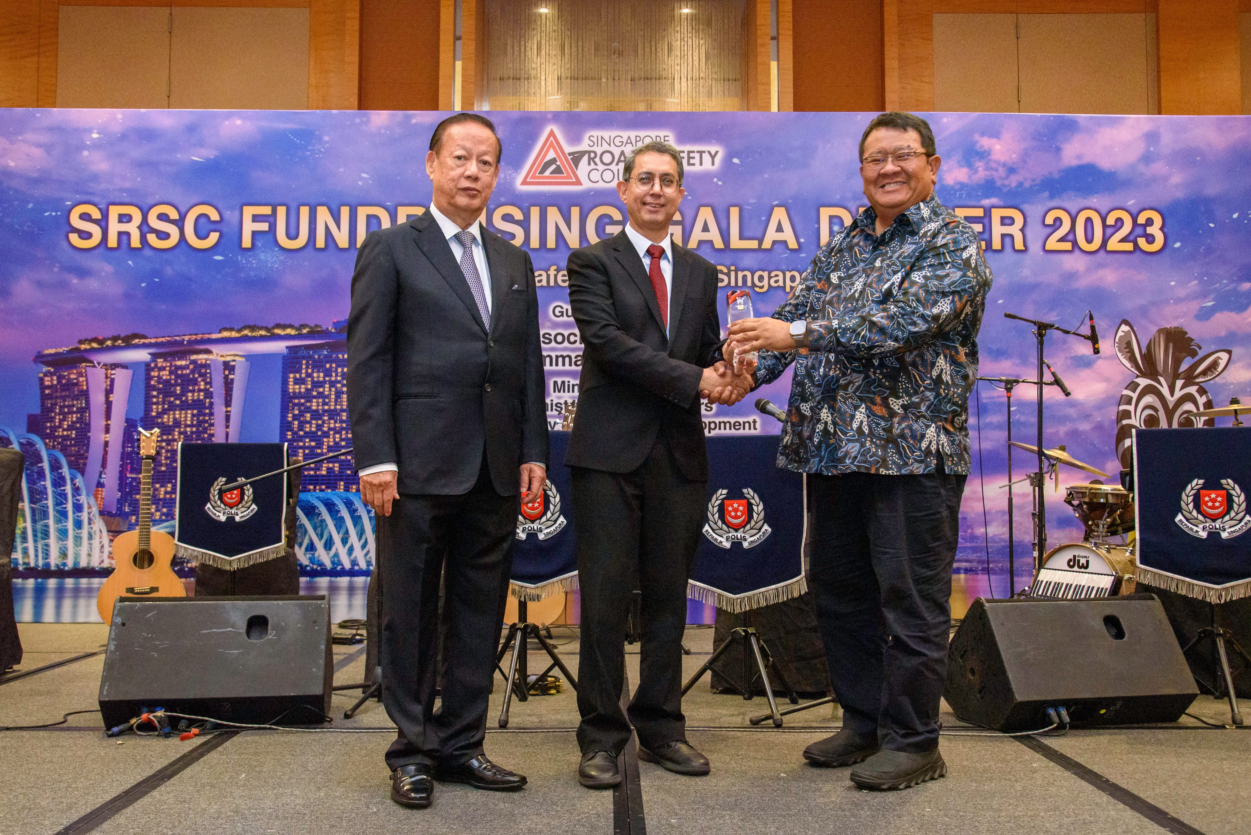 Three people stand in front of a backdrop at the SRSC Fundraising Gala Dinner 2023, with guitars and drums visible.