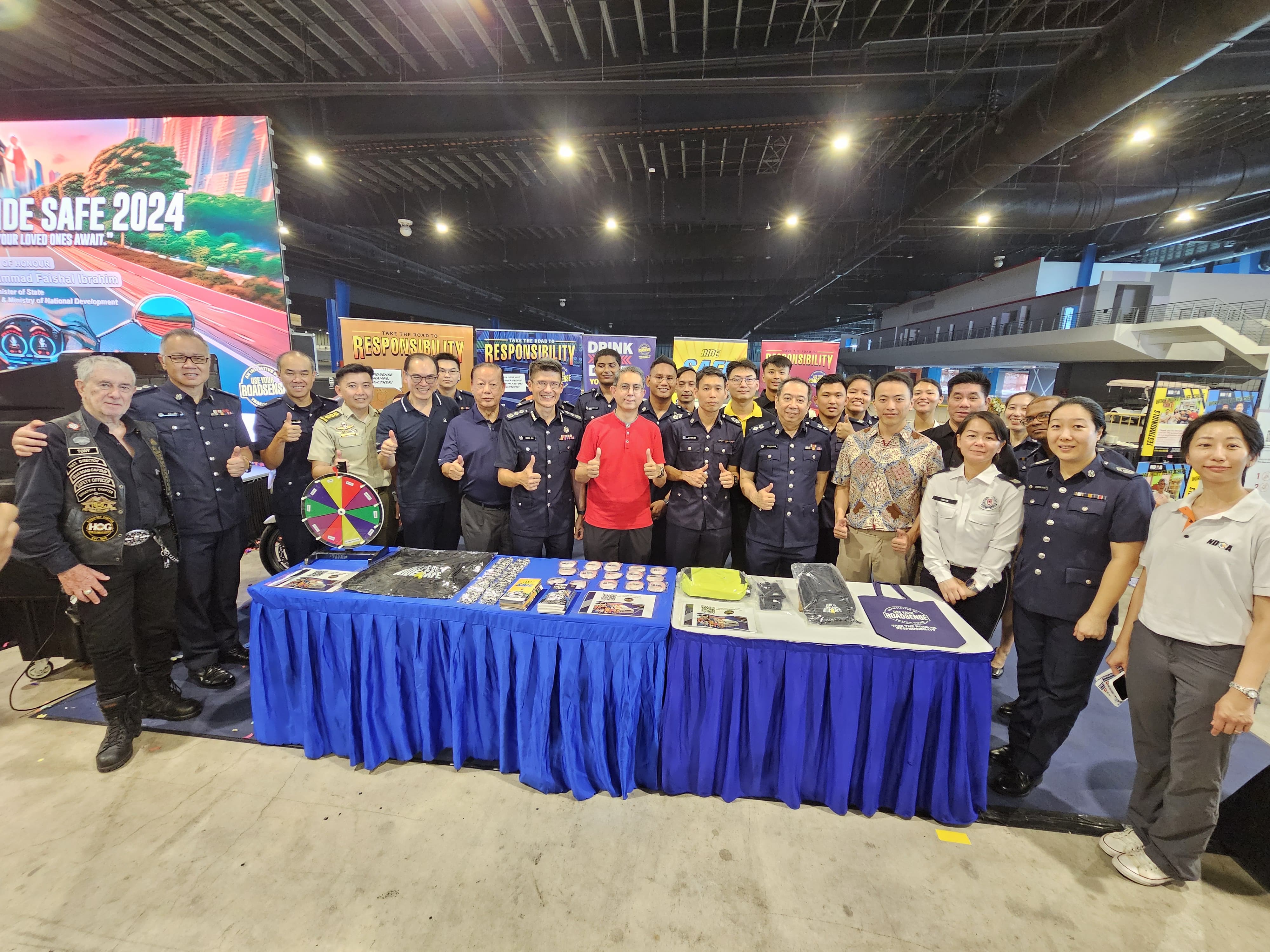 Group of people at an event with a table displaying various items and a large screen in the background.