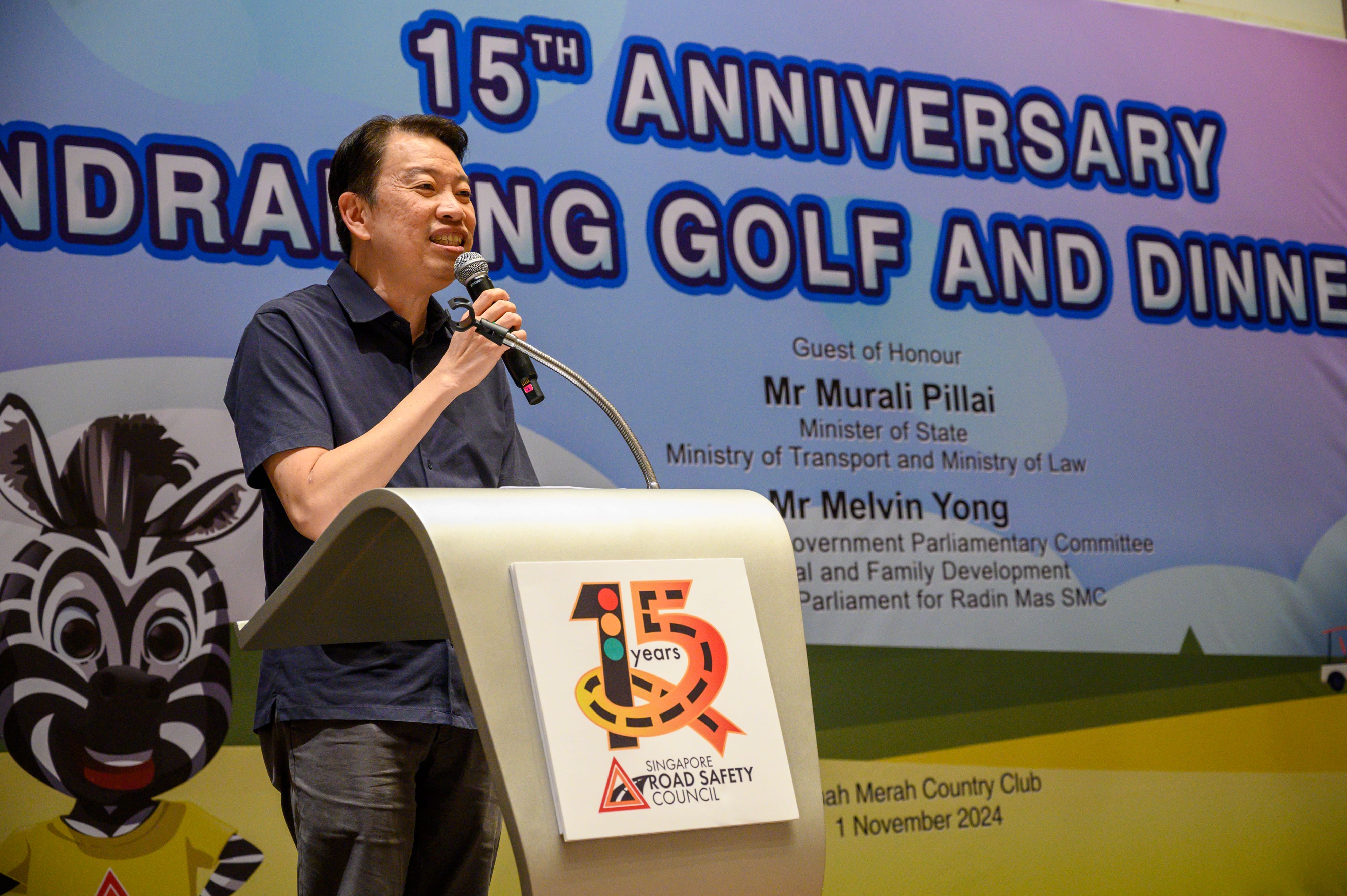 A person wearing a dark shirt speaks at a podium with a "15 years Singapore Road Safety Council" sign, in front of a banner.
