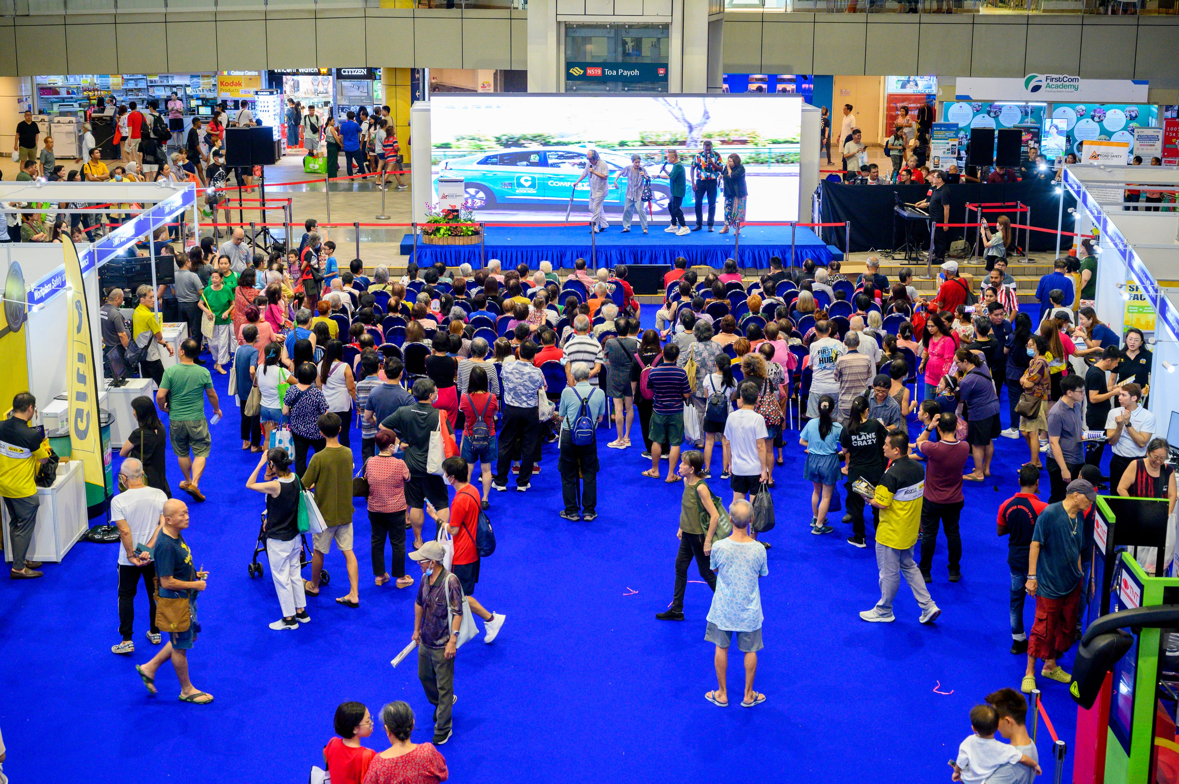 A large crowd in a convention center watches a stage presentation with a screen showing an image of a parked car.