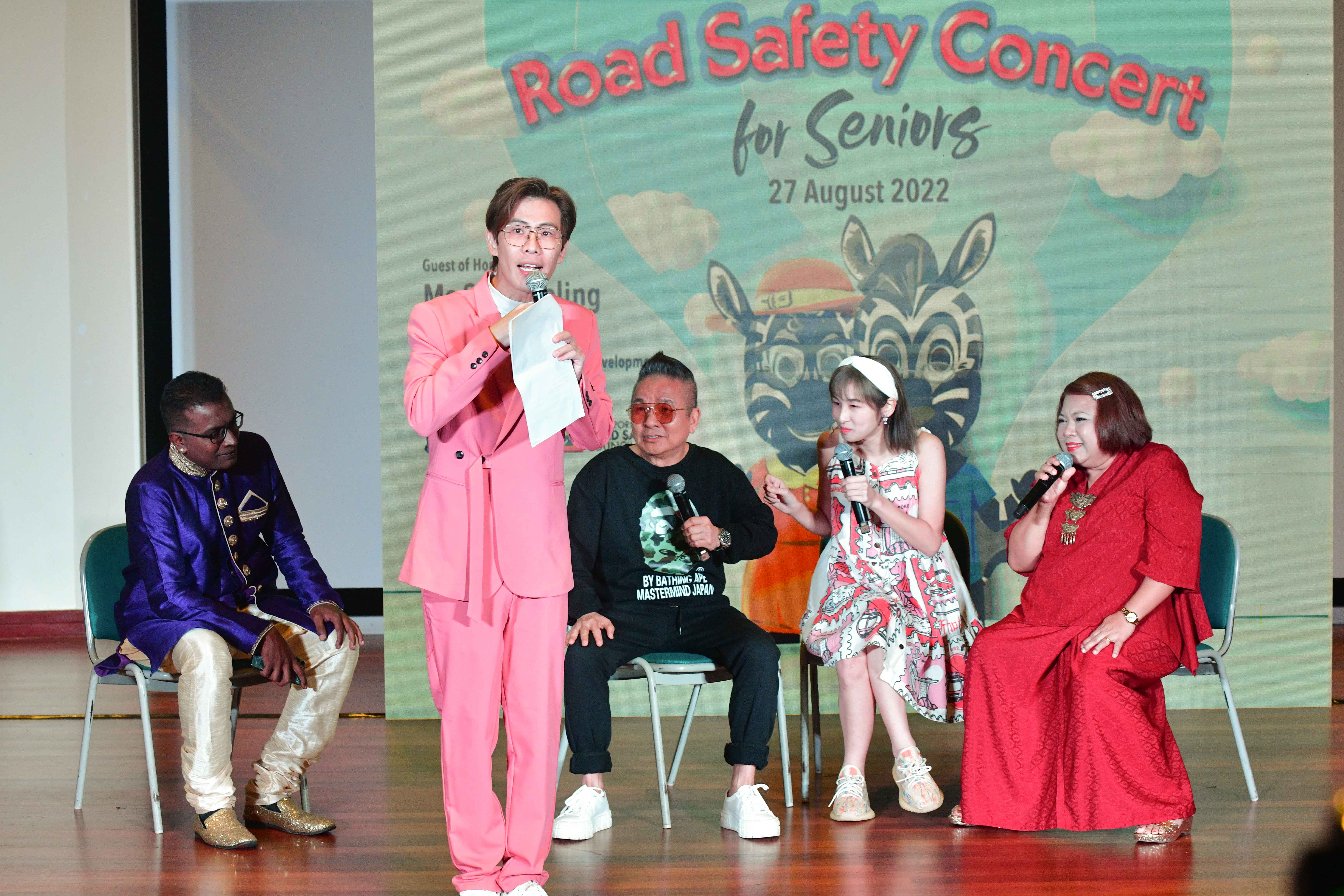 Five people are seated on stage holding microphones at a "Road Safety Concert for Seniors" event.