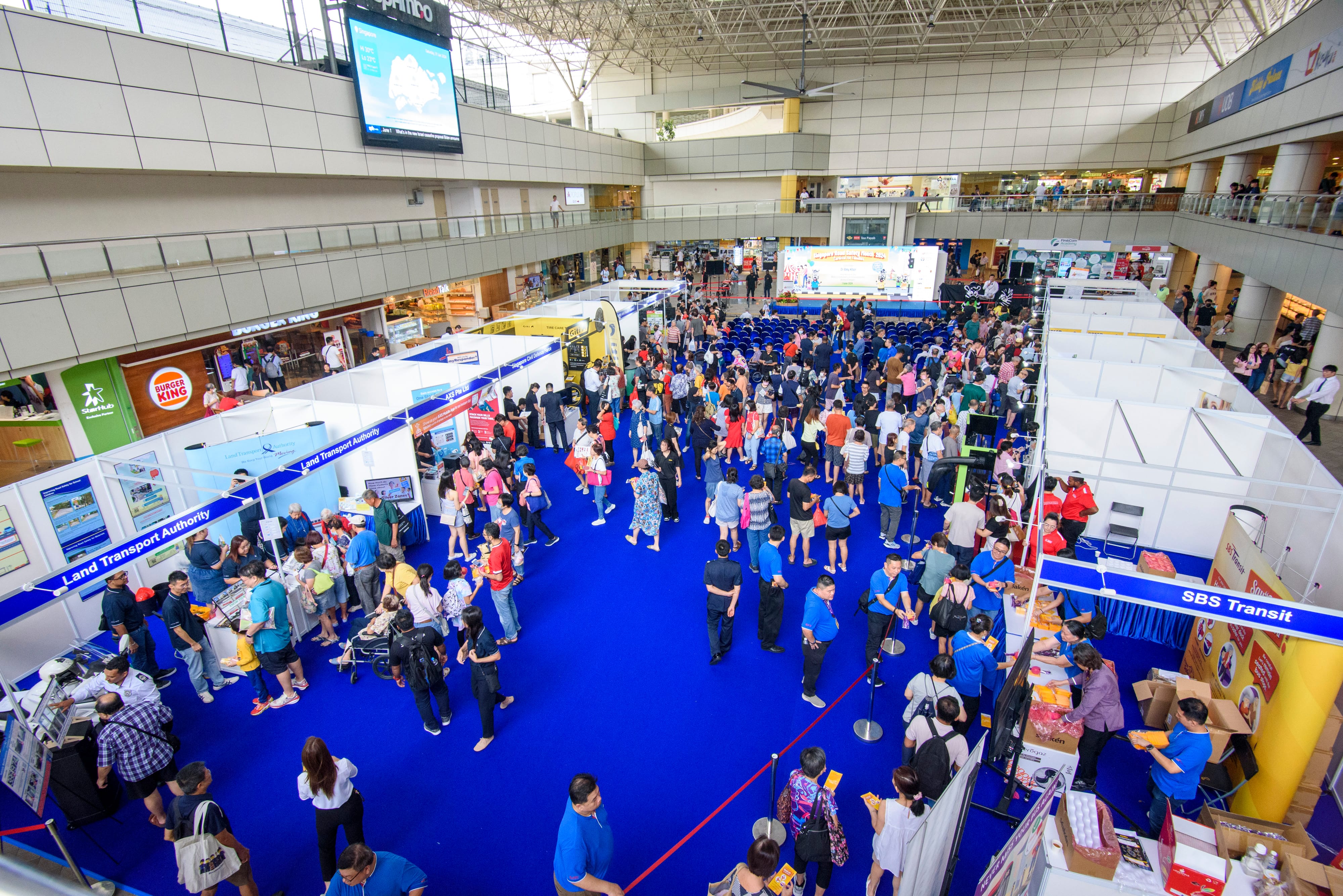 A crowded indoor exhibition hall with multiple booths, a stage with seating, and people walking on blue carpet.