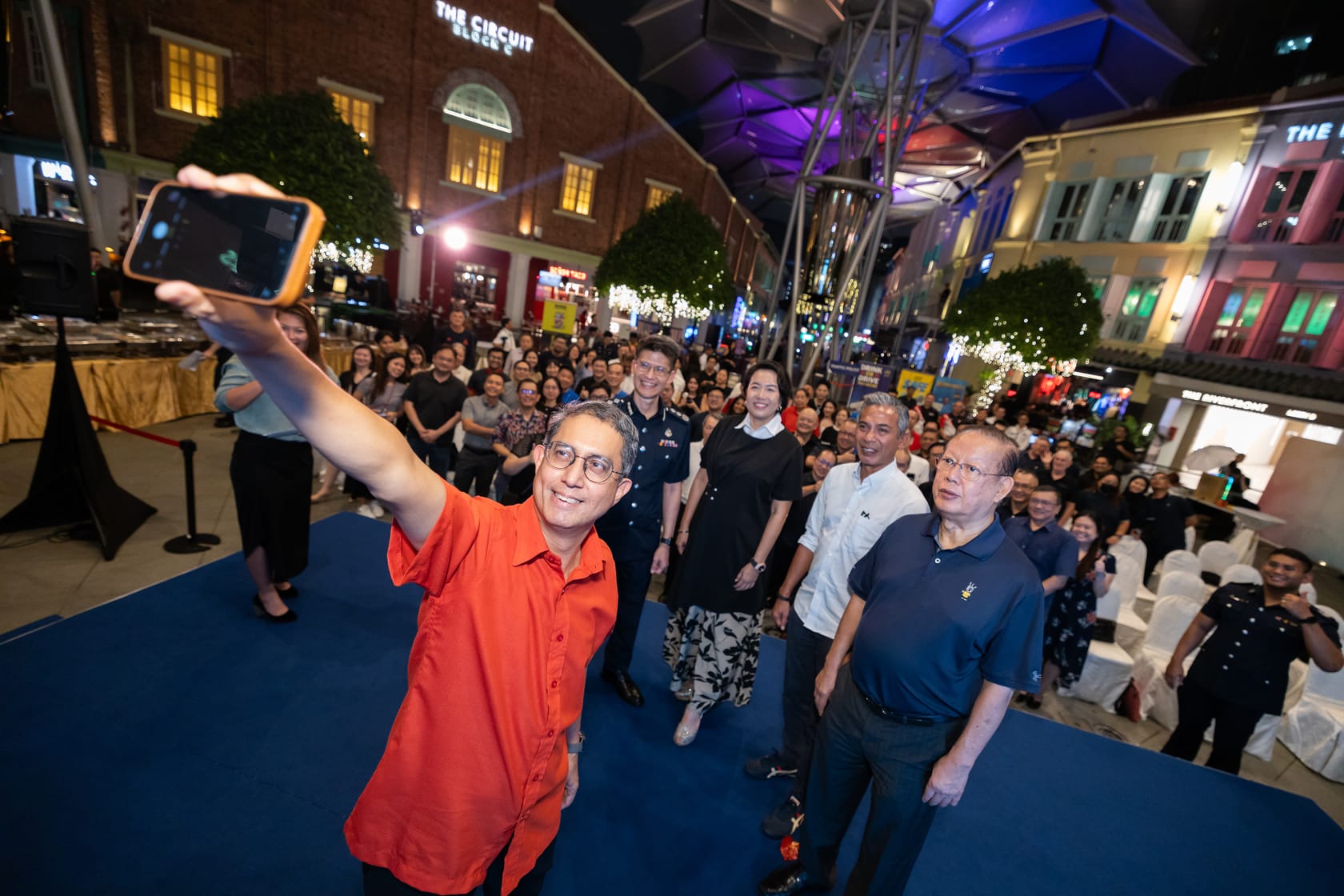 A person in a red shirt takes a selfie with a crowd behind them in a brightly lit outdoor area at night.