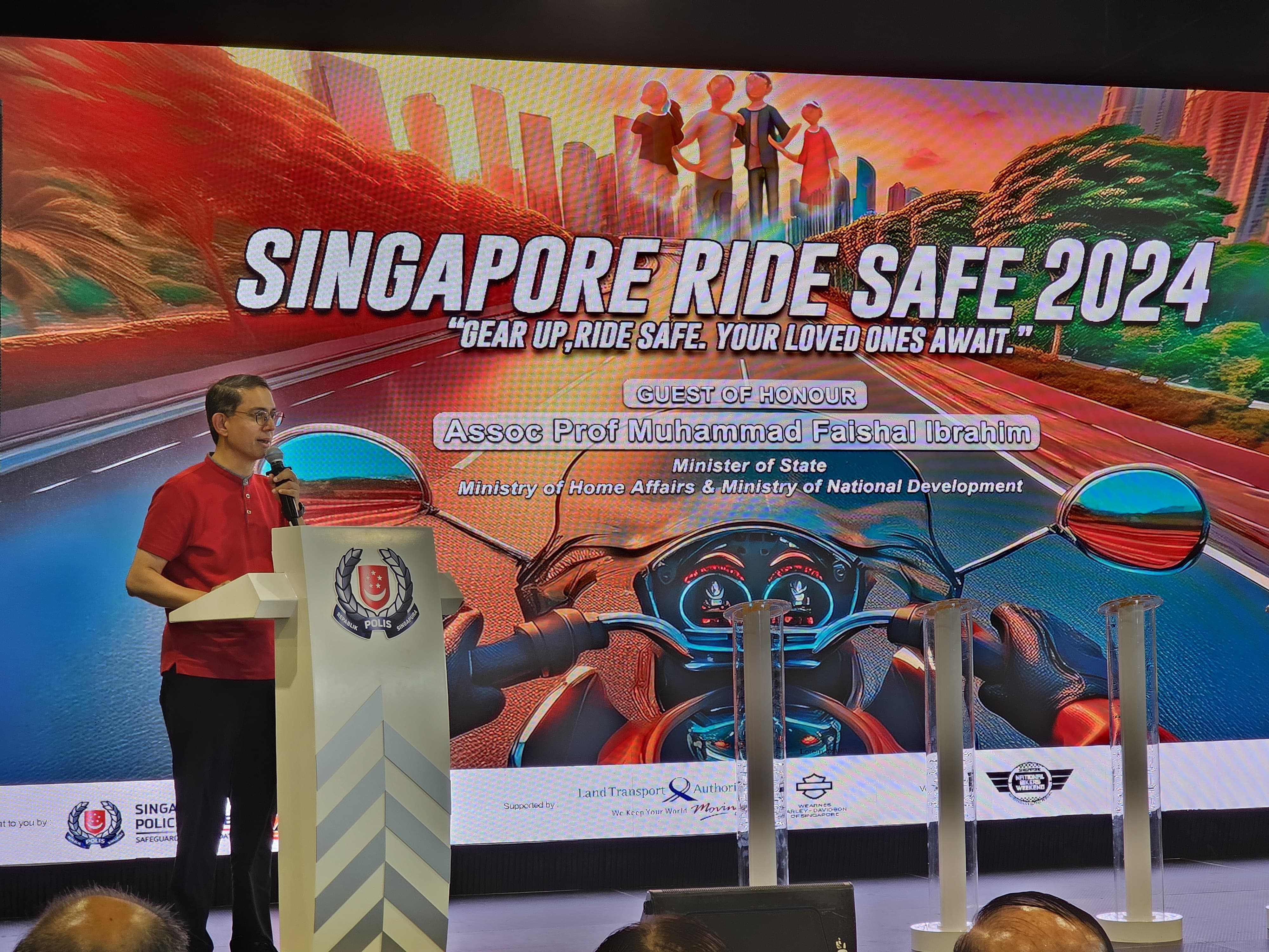 Man in red shirt speaks at a podium with a large backdrop promoting "Singapore Ride Safe 2024."