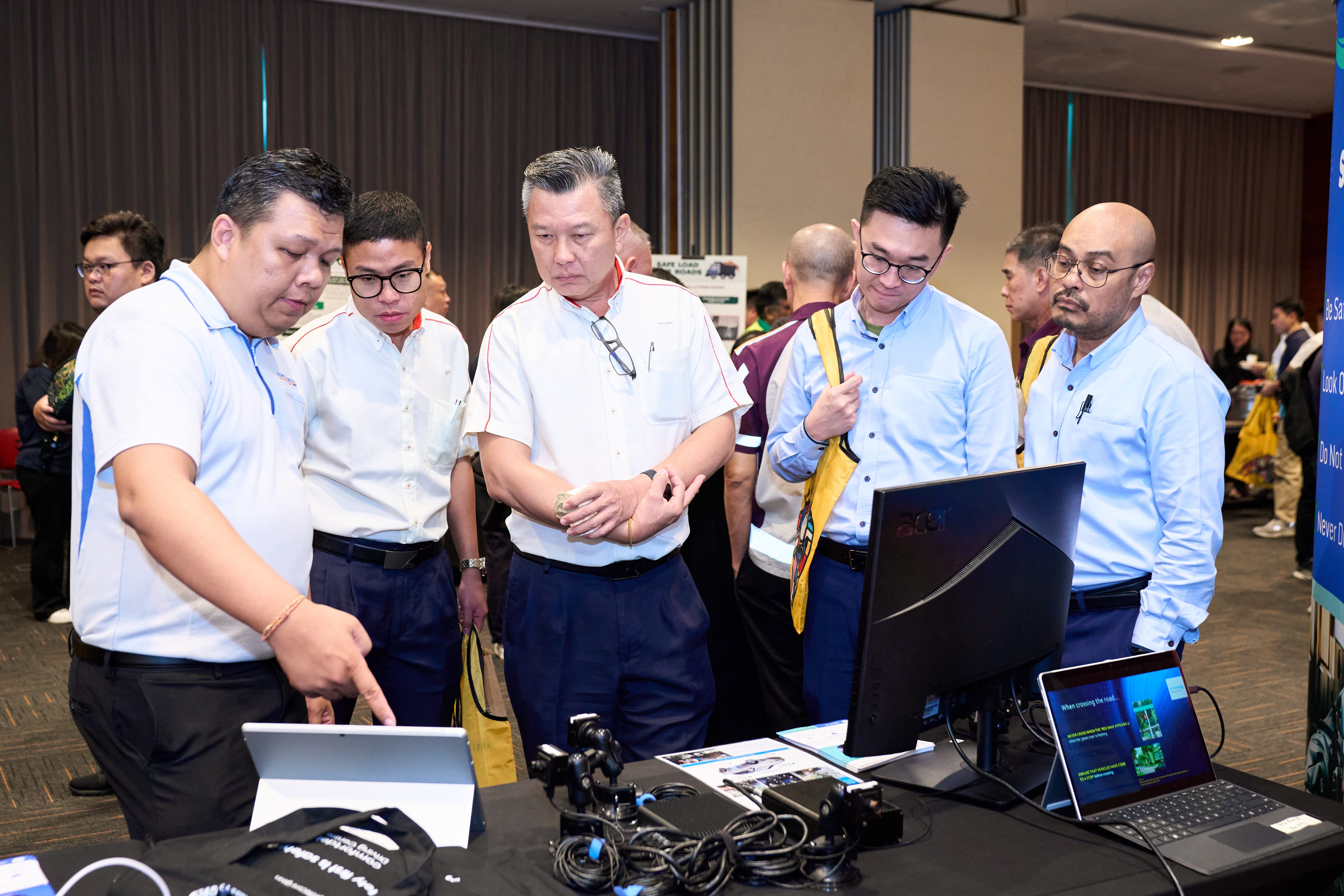 A group of people in white and blue shirts stand around a table with electronic equipment, looking at a laptop and monitor.